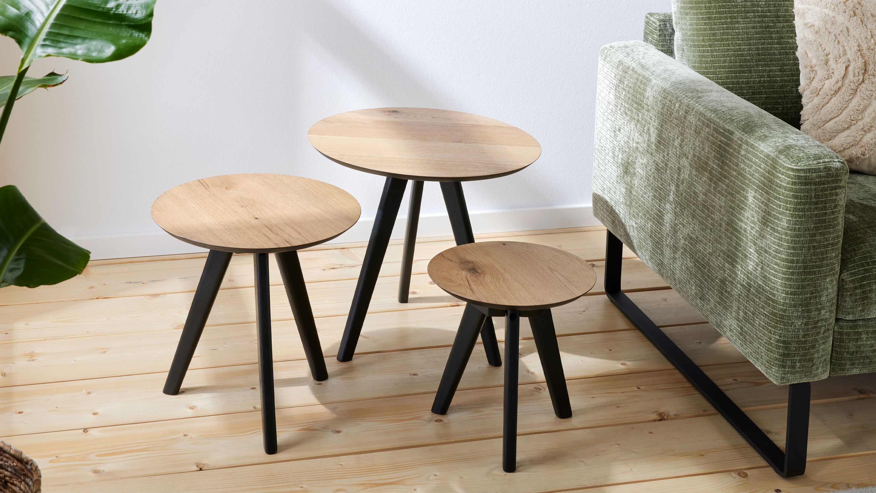 Dreiteiliges Couchtisch-Set mit runden hellen Tischplatten und schwarzen Beinen neben grünem Sofa