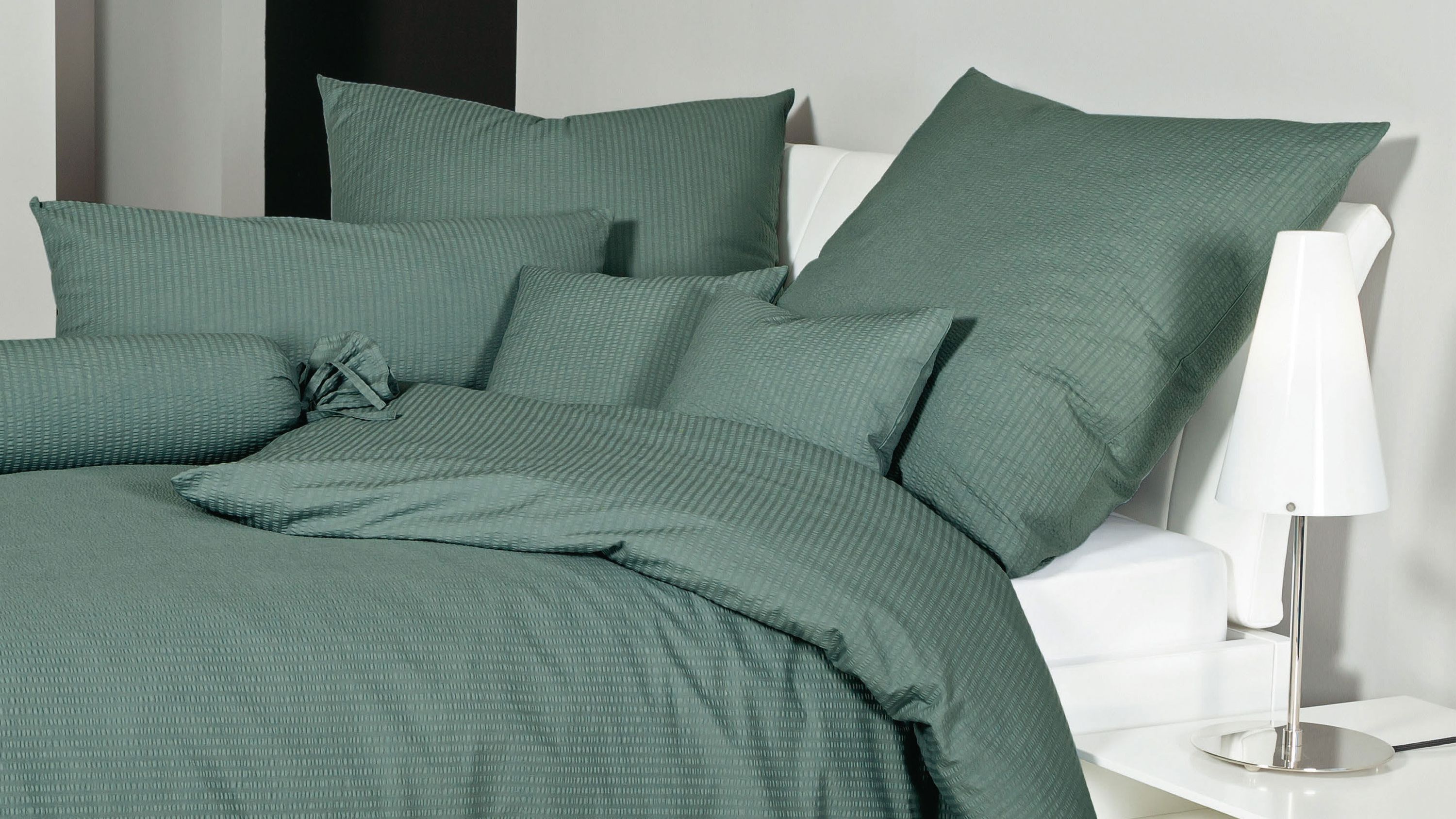 Salbeigrüne Bettwäsche mit Kissen und Decke auf weißem Bett mit weißer Lampe