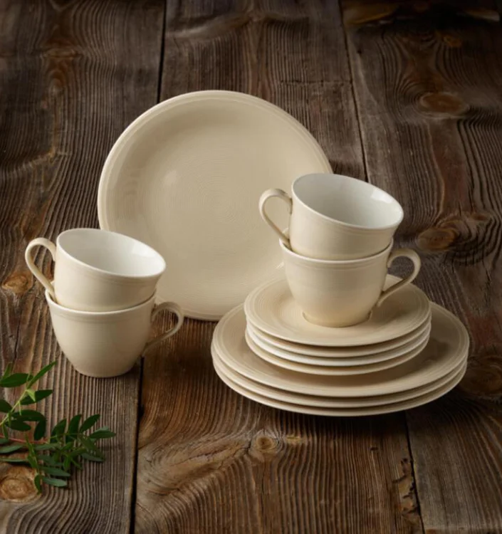 Cremefarbenes Keramikgeschirr-Set mit Tellern und Tassen auf rustikalem Holztisch mit grüner Pflanzendekoration.