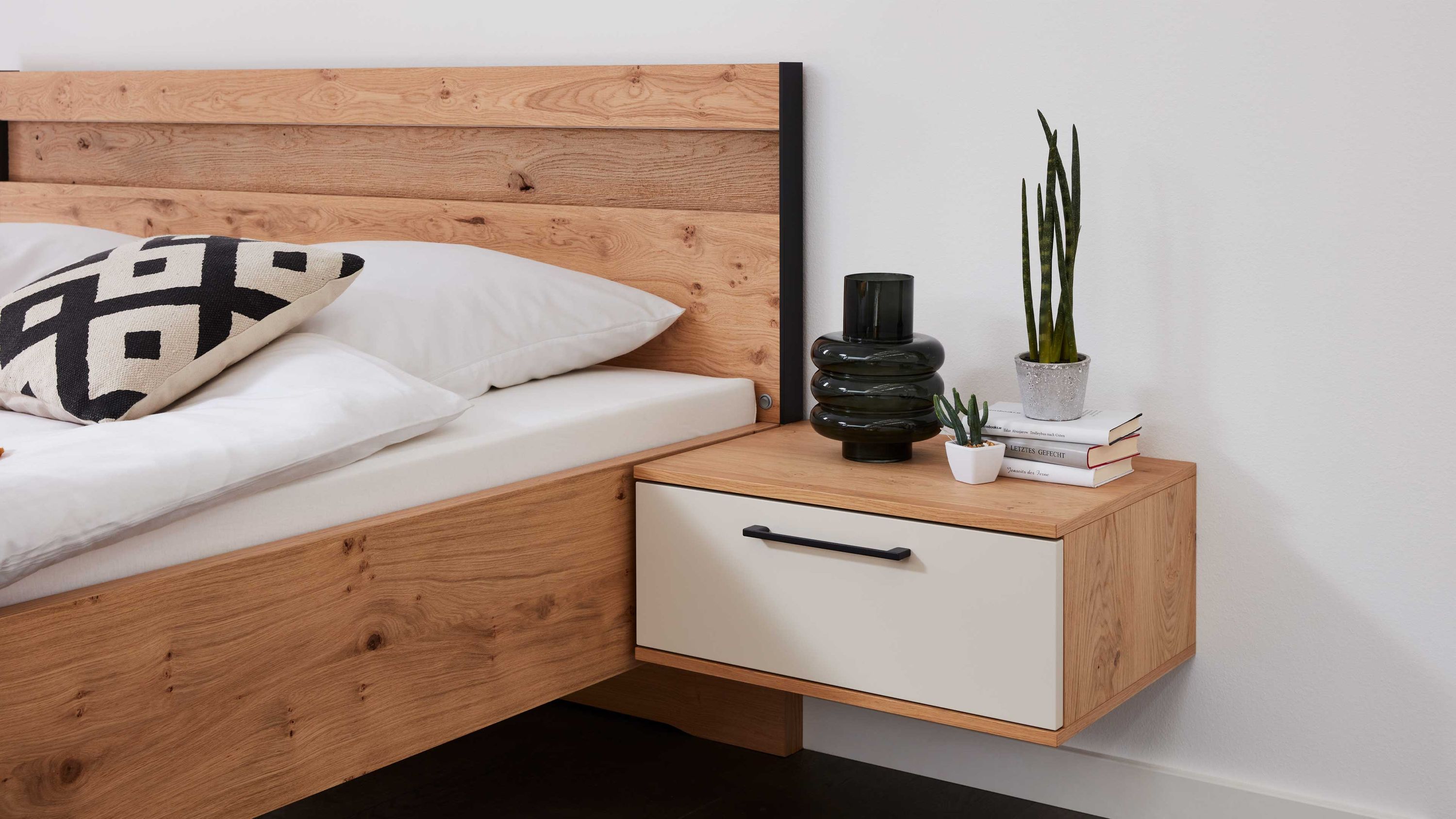 Schwebende Nachtkonsole mit einer weißen Schublade und Holzoptik in Sand und Balkeneiche neben einem Bett mit Holzrahmen