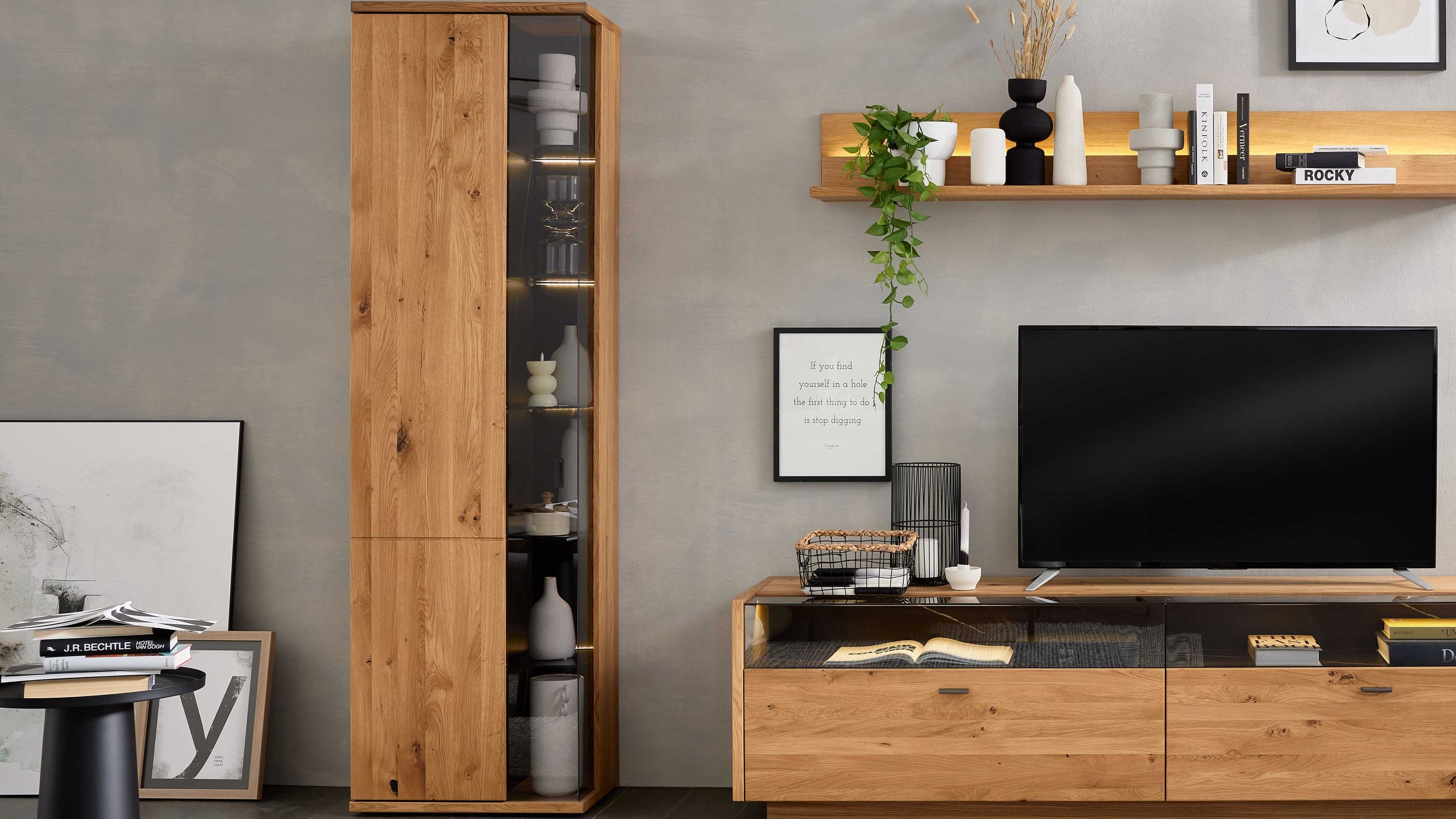 Hohe Vitrine mit einer Tür und Glasfach in hellem Holz Hohe Vitrine mit einer Holztür und einem Glasfach mit Beleuchtung in hellem Holz neben einem TV-Schrank