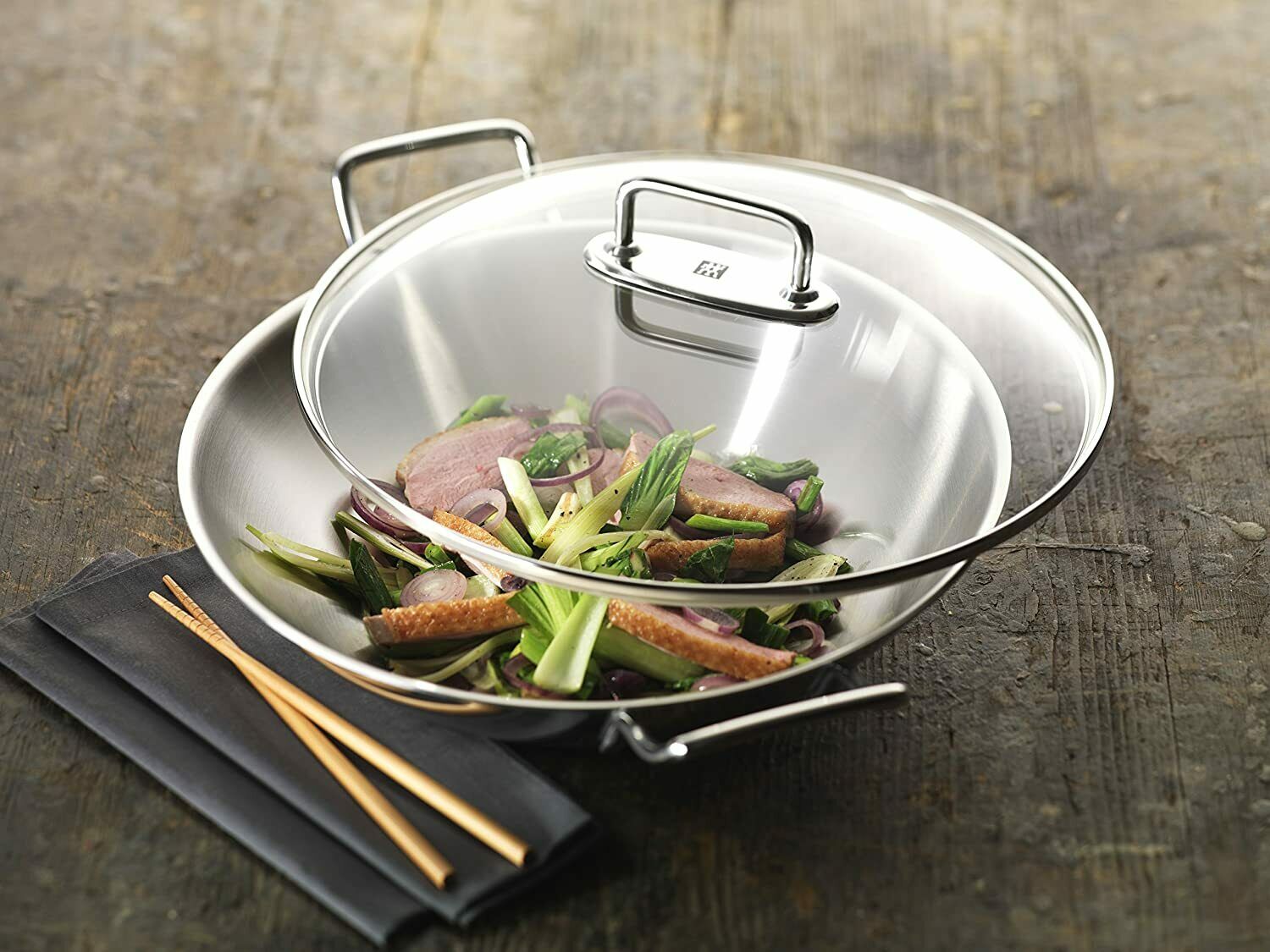 Edelstahl-Wok mit Gemüse und Fleisch auf schwarzer Unterlage, daneben Essstäbchen, frontal von oben fotografiert.