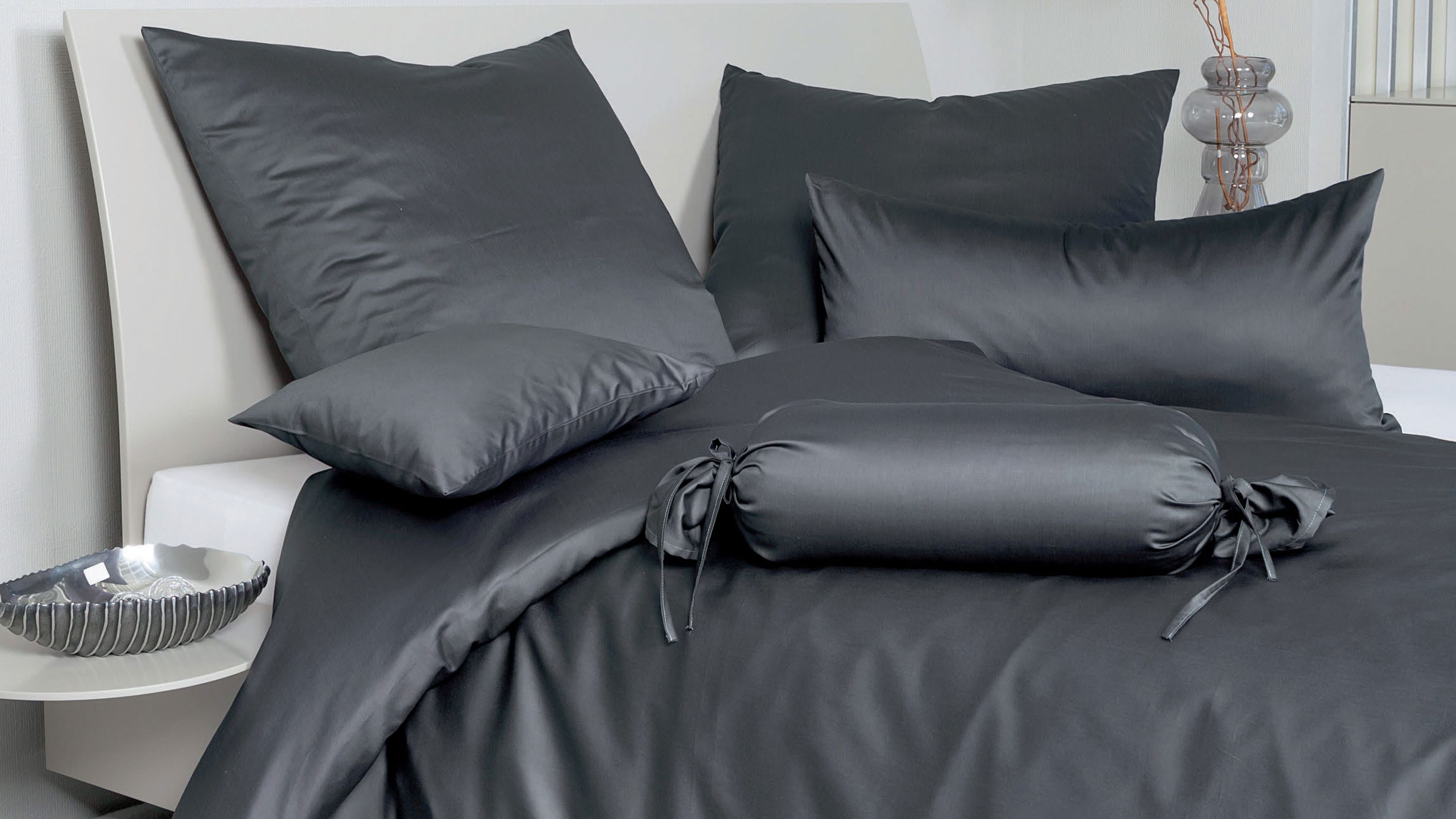 Dunkelgraue Bettwäsche mit Kissen und Zierrolle auf weißem Bett mit rundem Nachttisch