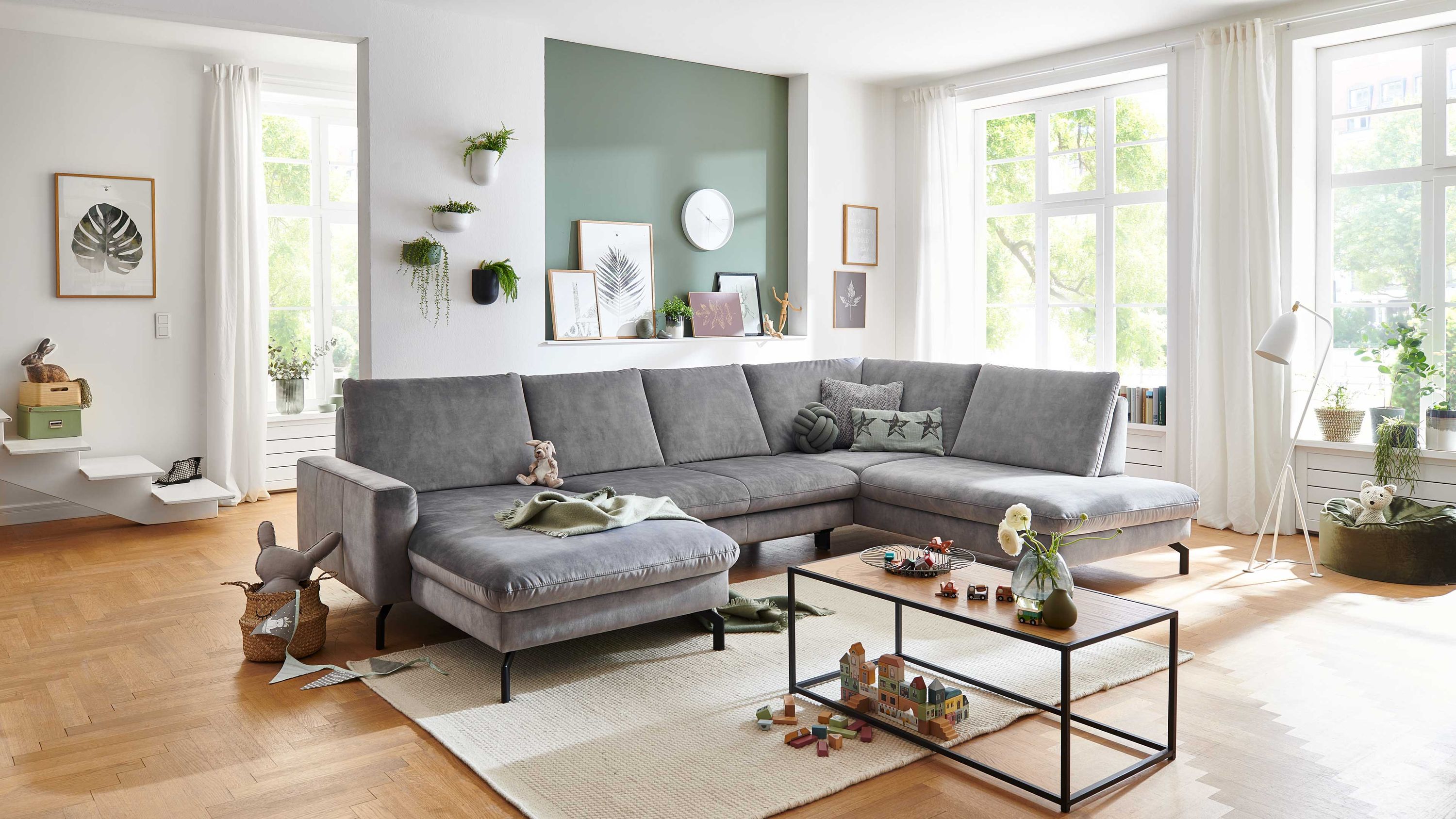 Großes silberfarbenes Velours Ecksofa mit schwarzen Beinen in hellem Wohnzimmer mit Holzboden