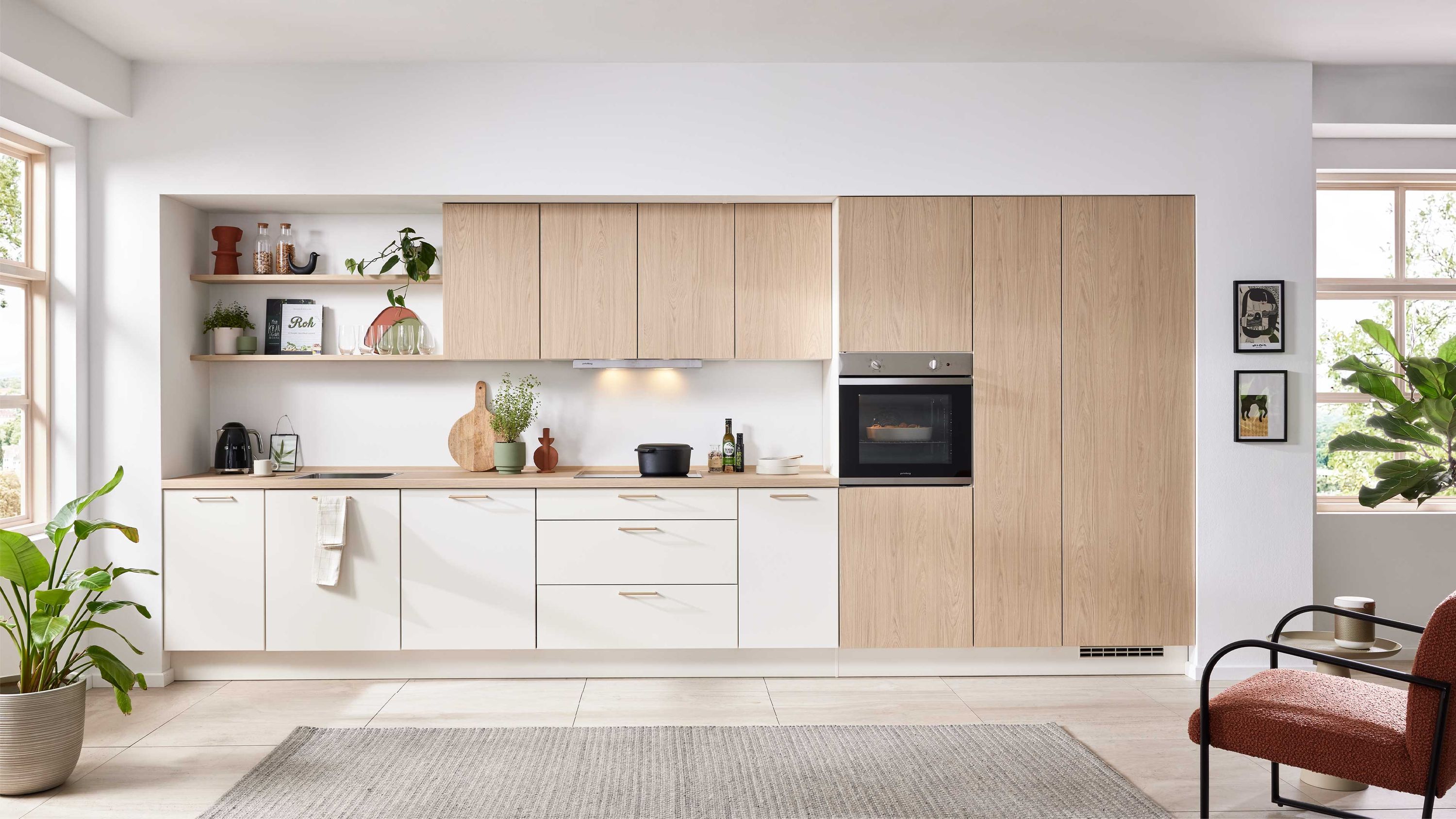 Helle Küche mit Holzoptik und Wohnbereich Weiße und Holzfronten offene Regale Einbauofen; Fensterlicht links Pflanze in Ecke Stuhl im Wohnbereich