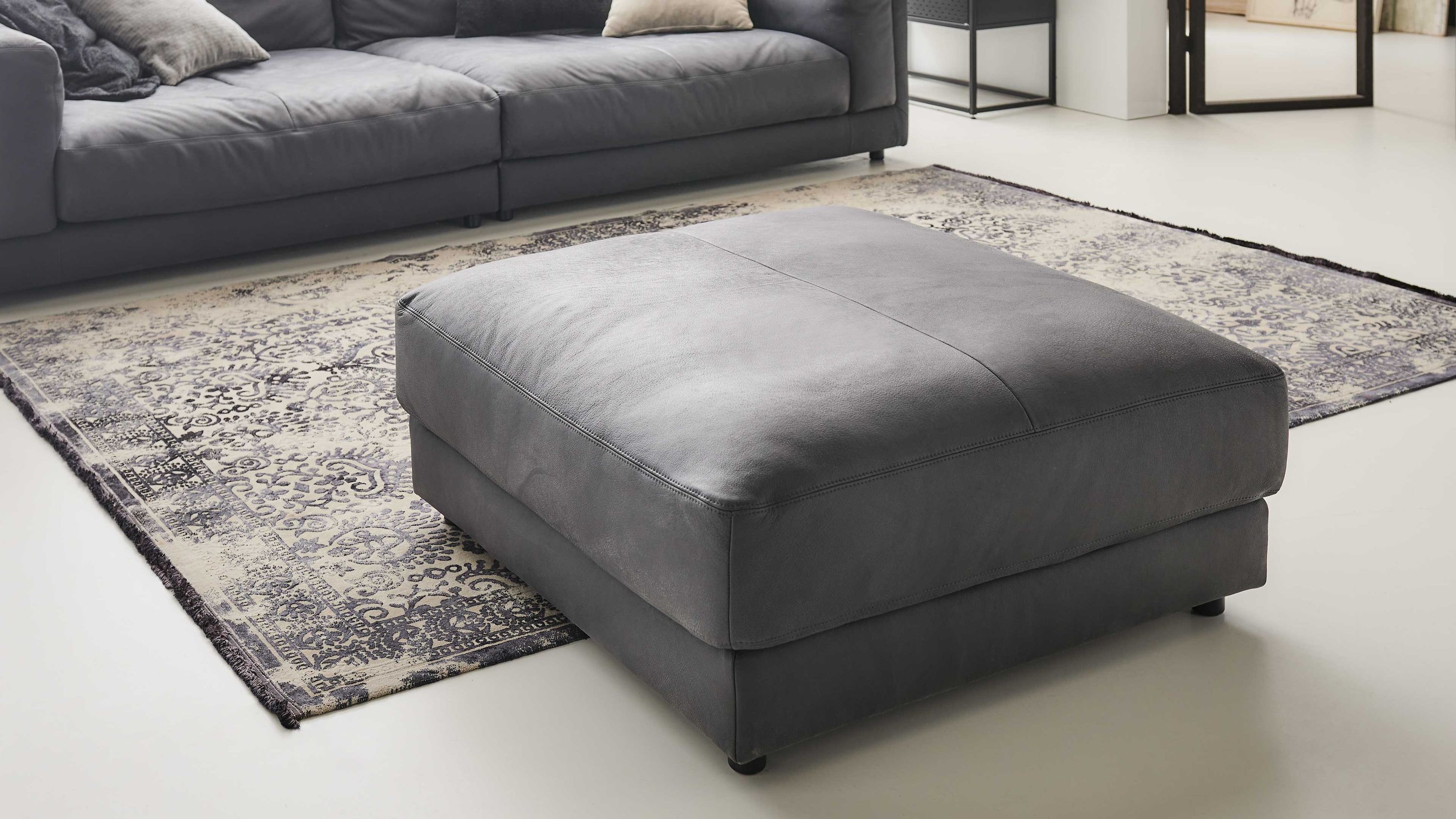 Quadratischer Hocker mit anthrazitfarbenem Lederbezug auf hellem Boden vor grauem Sofa