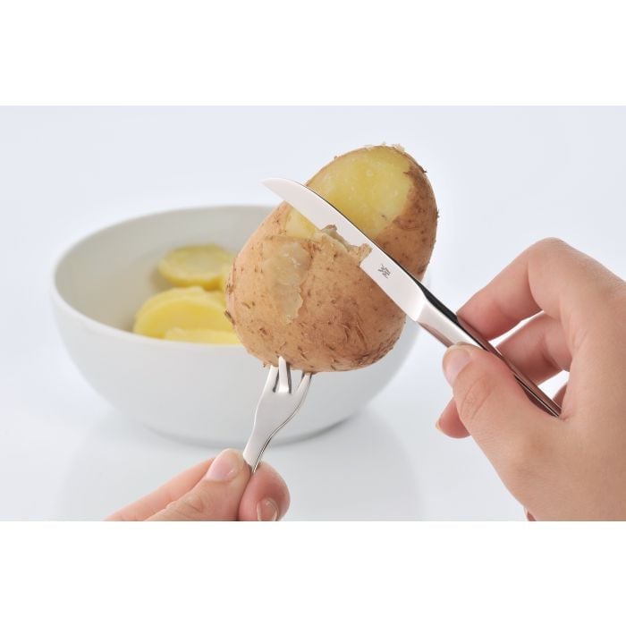 Hände schälen Kartoffel mit Pellkartoffelgabel und Schälmesser vor weißer Schale mit Kartoffelscheiben, Nahaufnahme