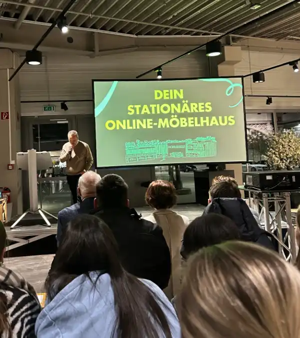 Prokurist Tobias Lehr spricht mit Mikrofon vor Publikum, hinter ihm eine Leinwand mit der Aufschrift „Dein stationäres Online-Möbelhaus“.