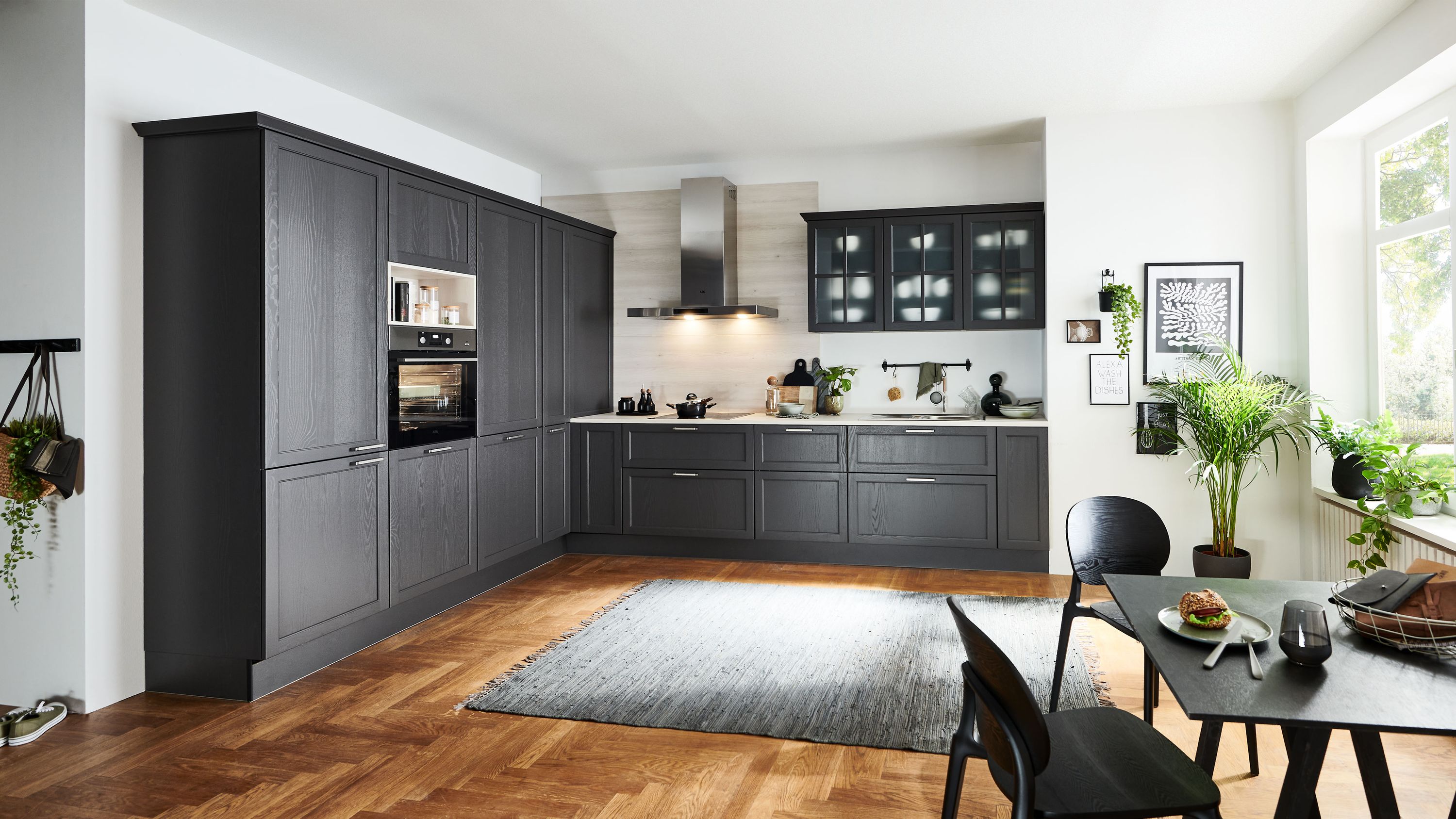Dunkelgraue L-Küche im Landhausstil mit Glashängeschränken Einbaugeräten Holzarbeitsplatte und hellem Fensterbereich.