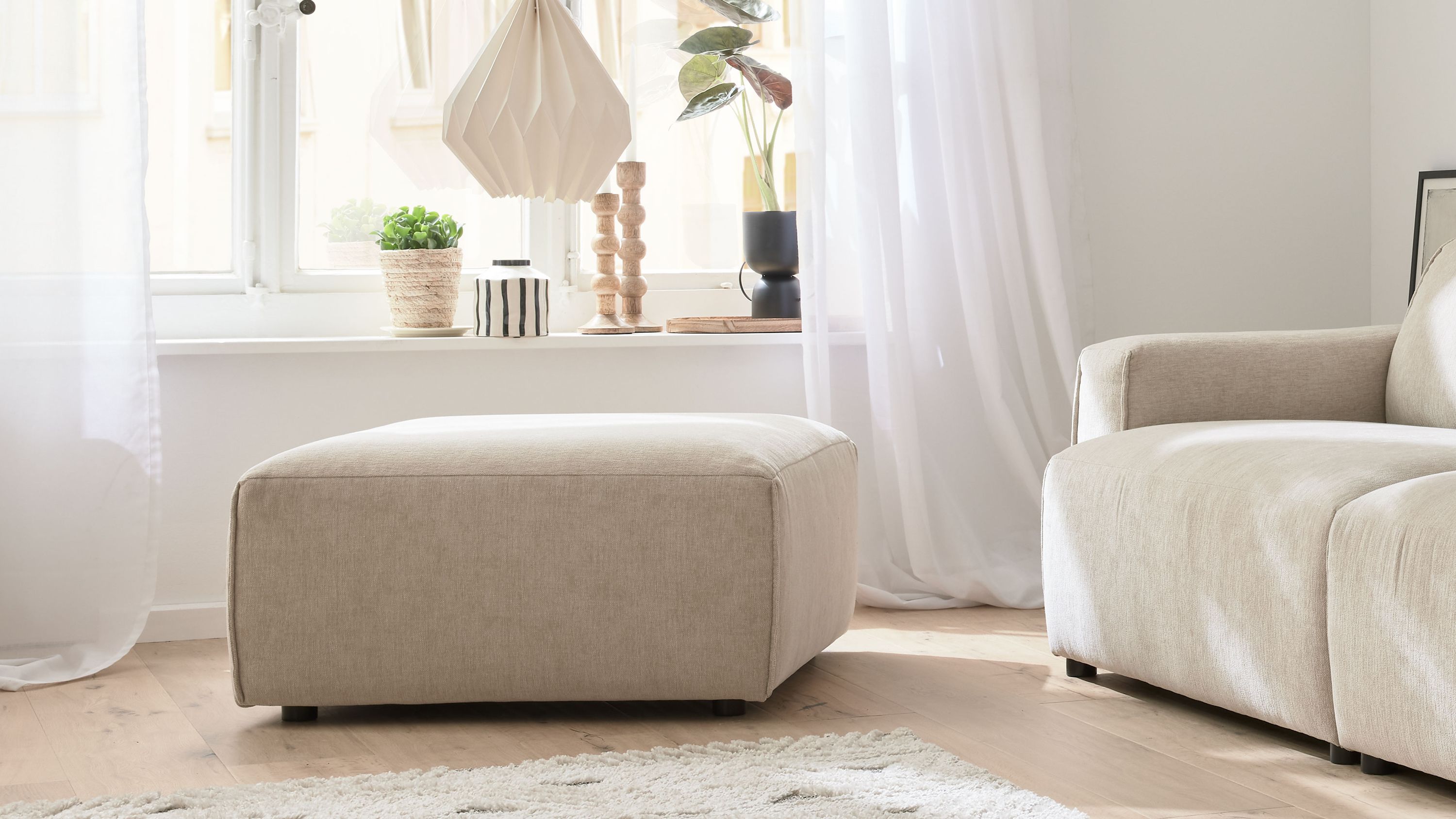 Quadratischer Polsterhocker mit champagnerfarbenem Bezug in einem hellen Wohnzimmer neben einem Sofa