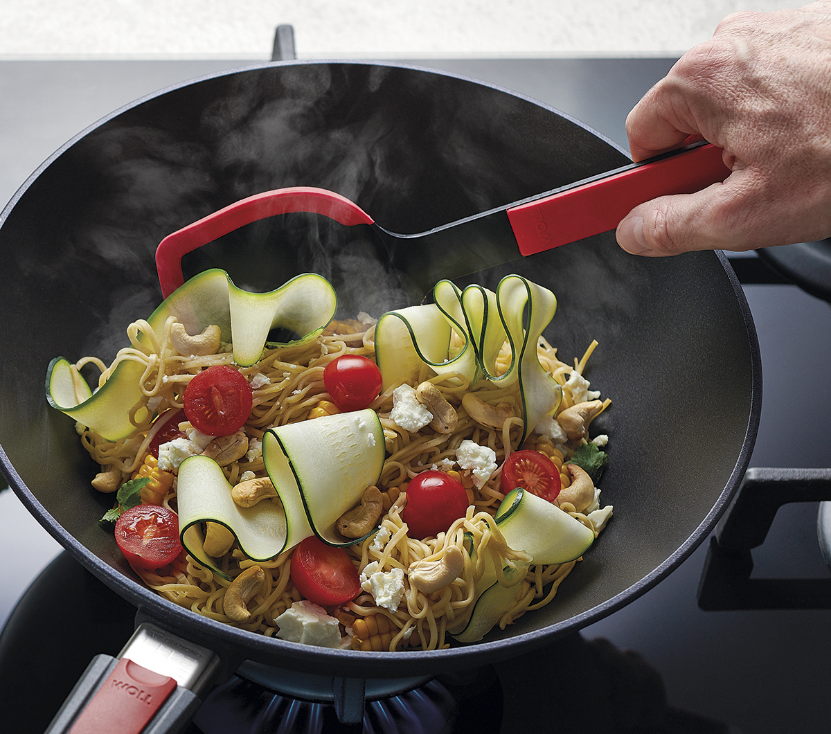 Schwarzer Wok auf Herd Hand mit rotem Wokwender rührt Zucchini und Cherrytomaten leicht von oben aufgenommen