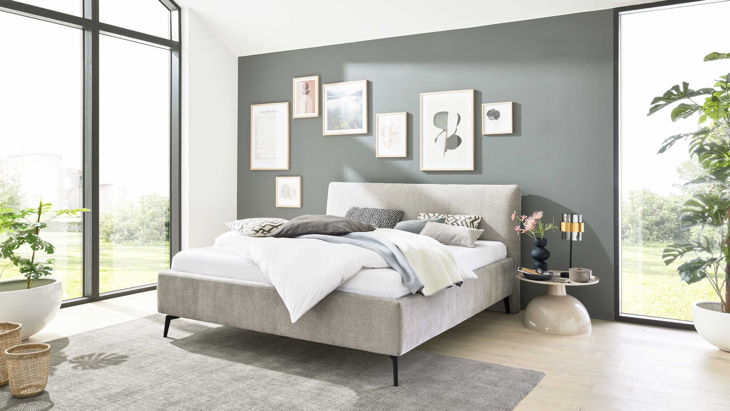 Polsterbett mit silberfarbenem Bezug und Metallgestell in modernem Schlafzimmer mit grauer Wand und großen Fenstern.