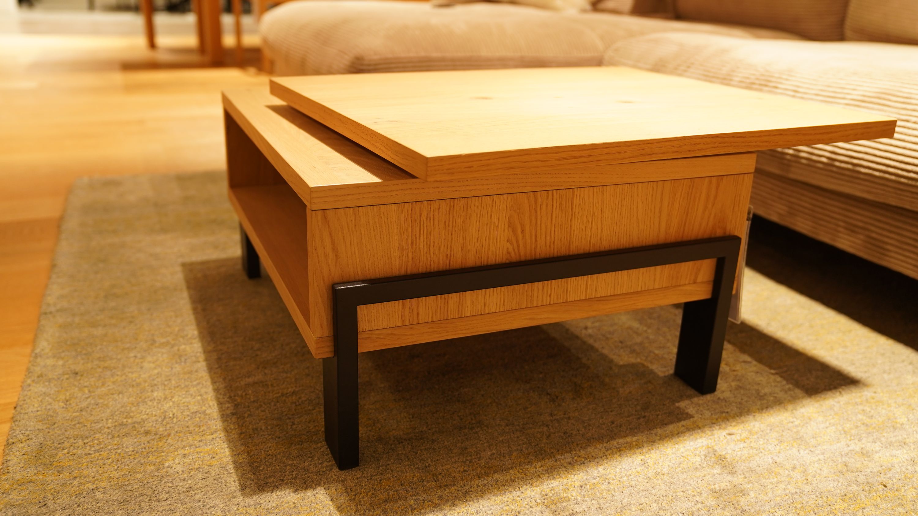 Holz-Couchtisch mit versetzter Platte, schwarzem Gestell und offenem Fach auf Teppich vor Sofa. Quadratischer Couchtisch mit versetzter Tischplatte und Stauraumfach