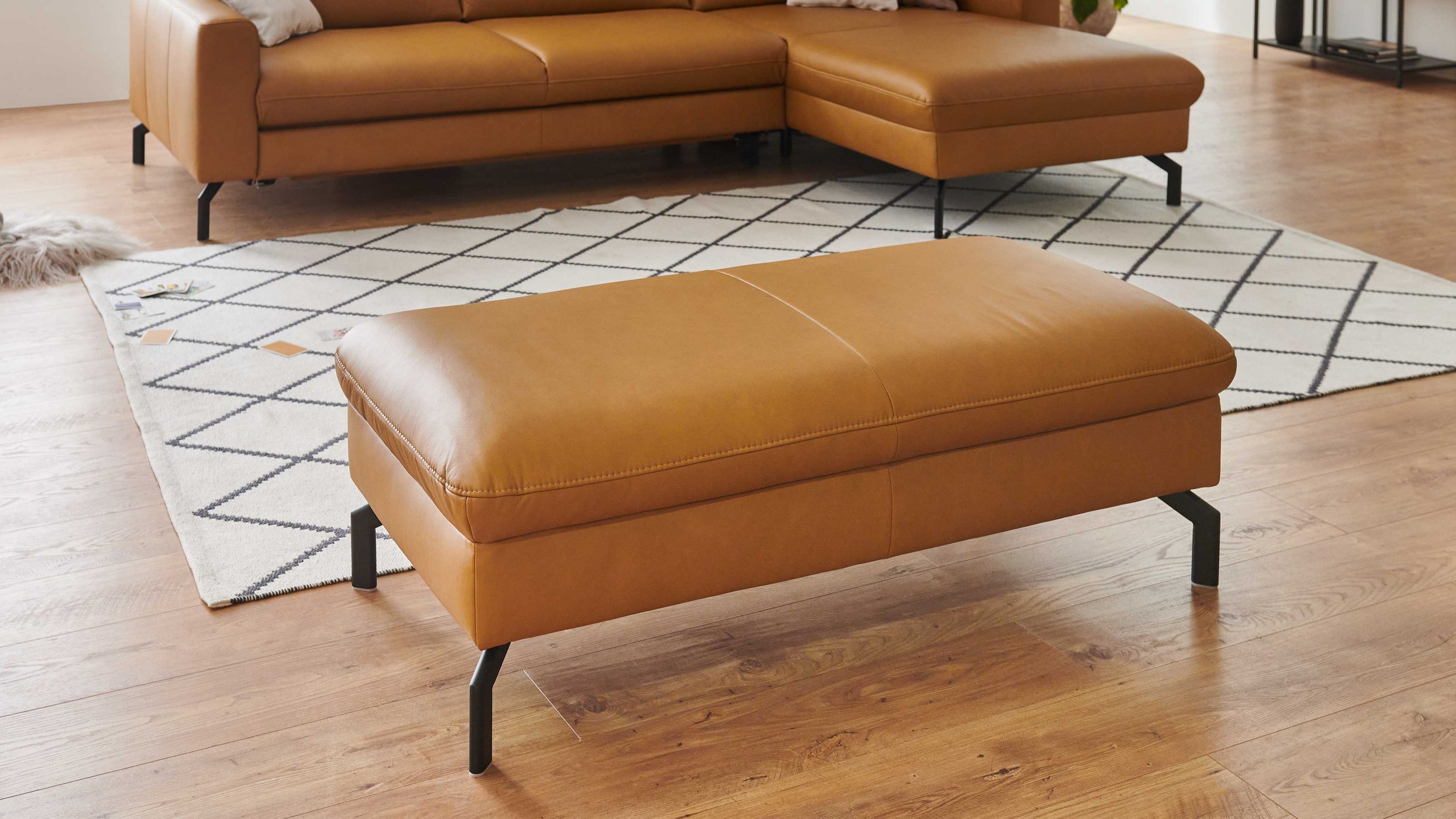 Rechteckiger Hocker mit kurkumafarbenem Lederbezug und schwarzen Metallfüßen vor braunem Ecksofa auf hellem Teppich