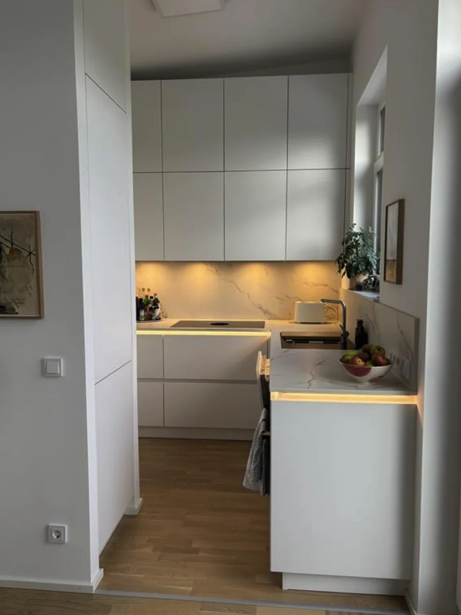 Kompakte weiße Küche mit Hochschränken, Holzarbeitsplatte, Spüle, Backofen und warmer Unterbauleuchtung.