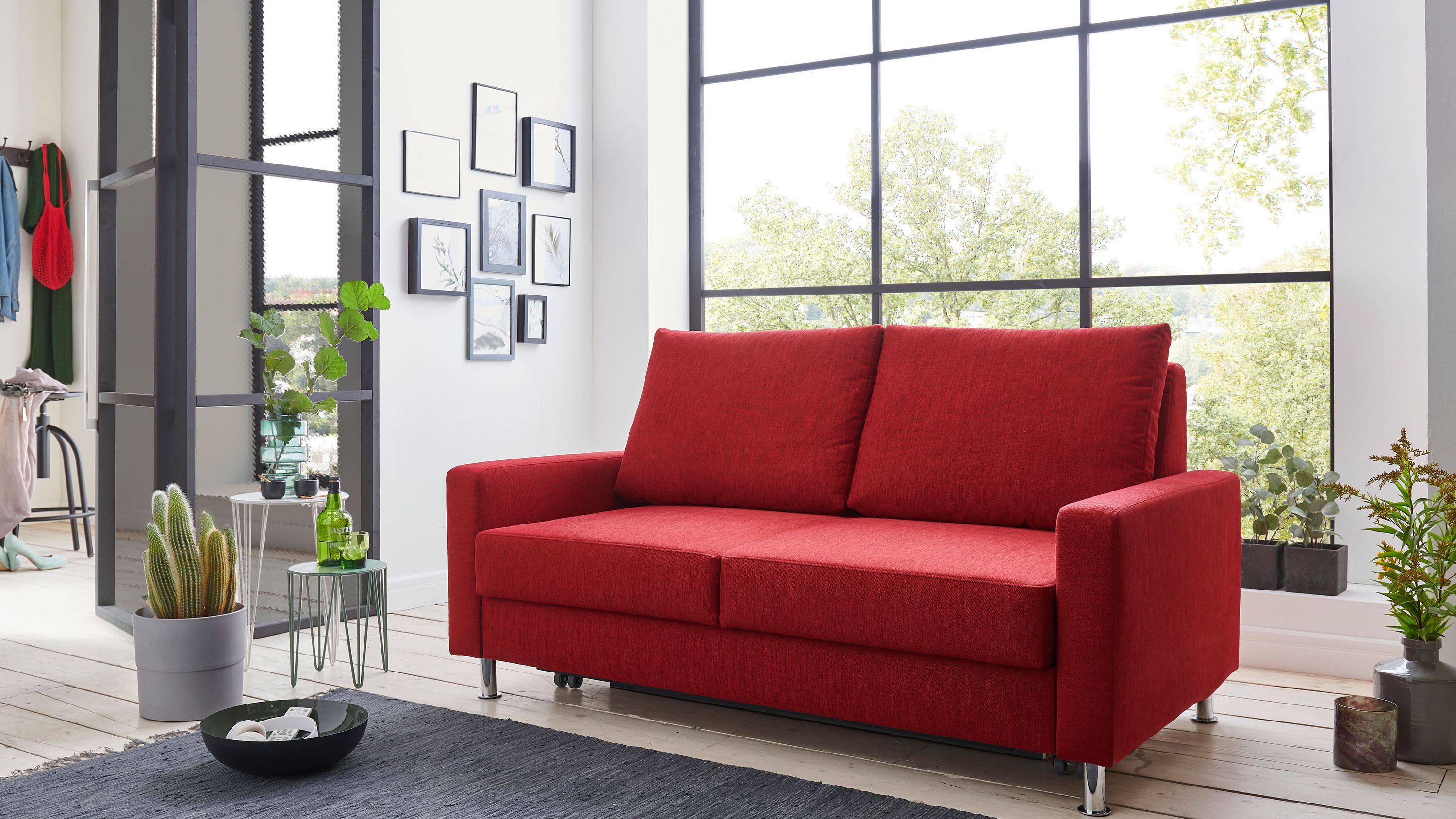 Rotes Schlafsofa mit zwei Rückenpolstern und Armlehnen in modernem Wohnzimmer vor großem Fenster