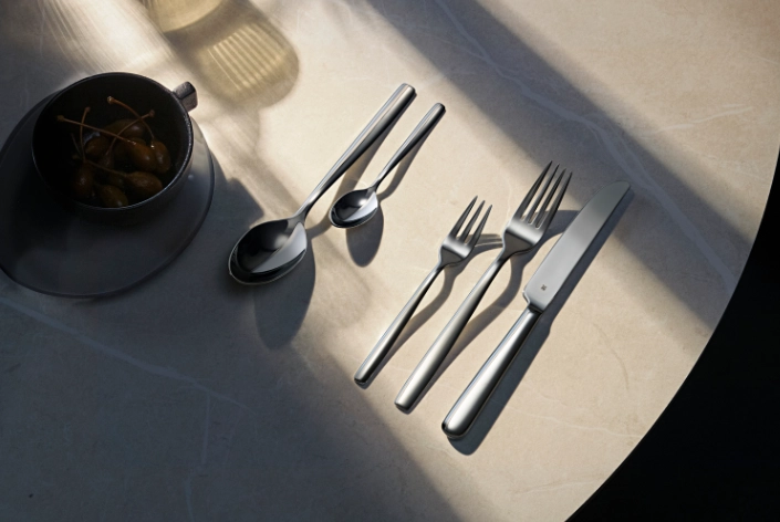 Besteck WMF2 Modernes Edelstahl-Besteckset mit Messer, Gabeln und Löffeln auf hell beleuchtetem Tisch neben einer Schale mit Oliven.