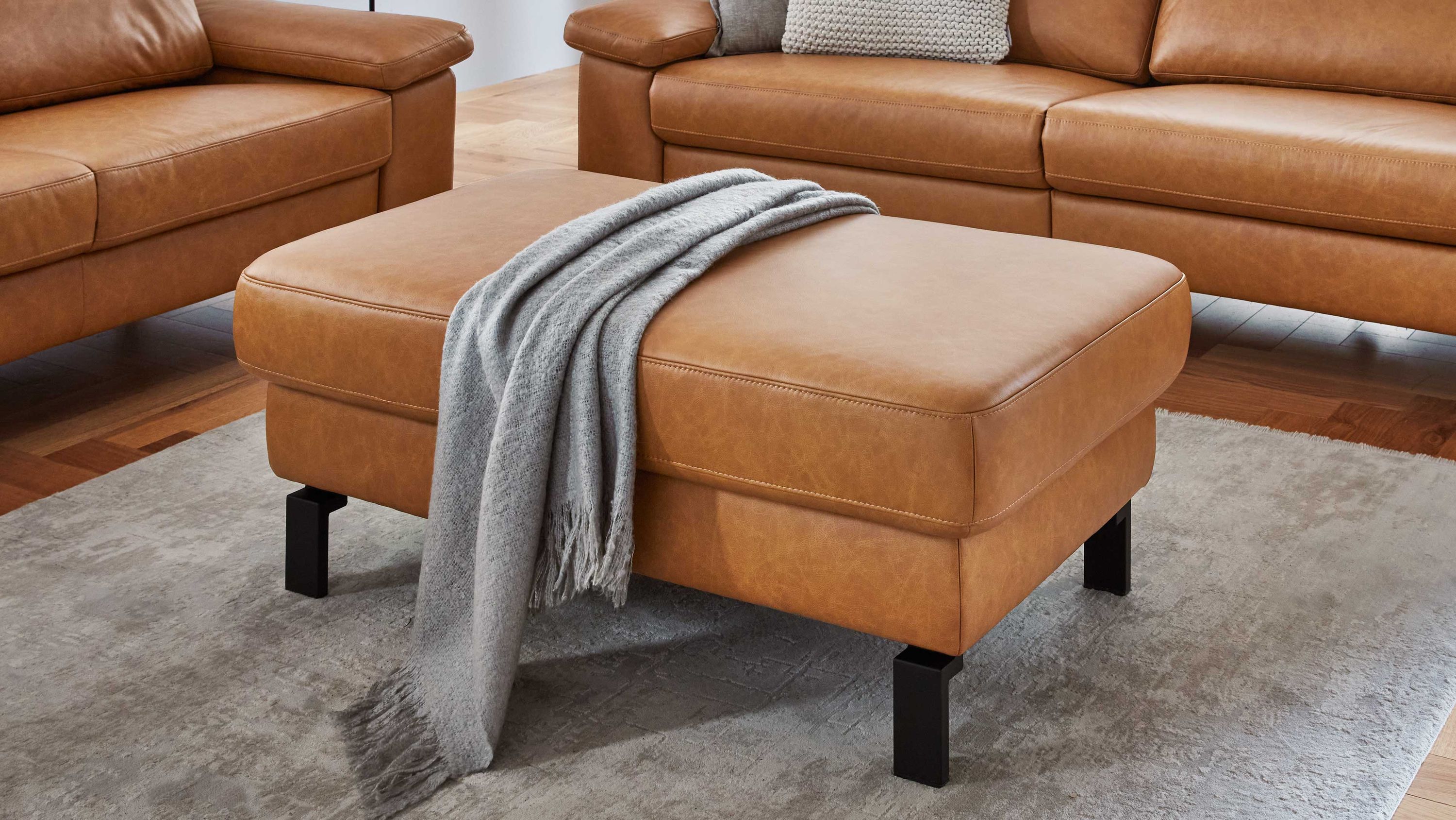 XXL-Hocker mit Lederbezug und Metallfüßen Rechteckiger Hocker mit cognacfarbenem Lederbezug und schwarzen Metallfüßen auf grauem Teppich