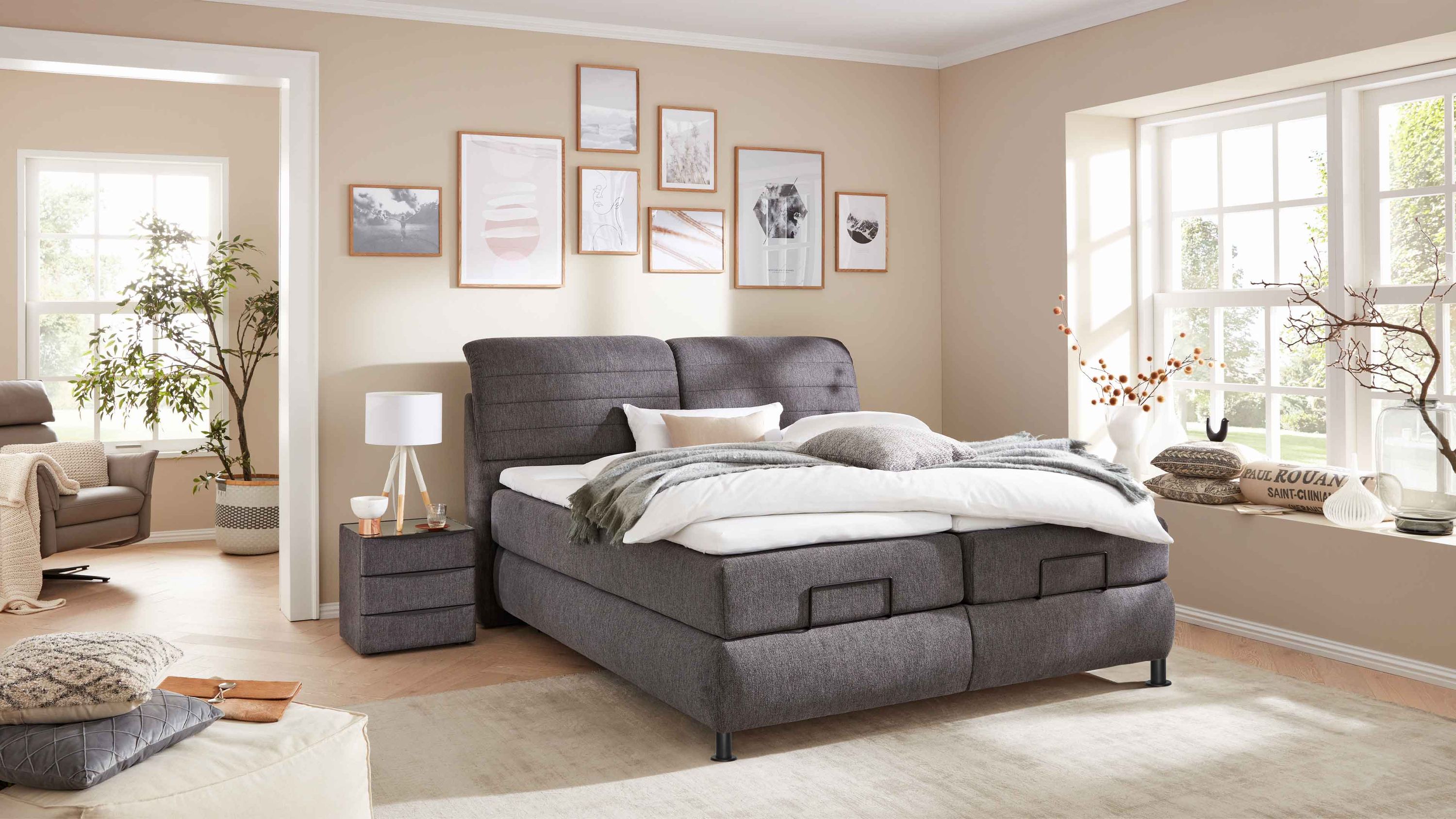 Boxspringbett mit anthrazitfarbenem Stoffbezug und gepolstertem Kopfteil in einem hellen Schlafzimmer