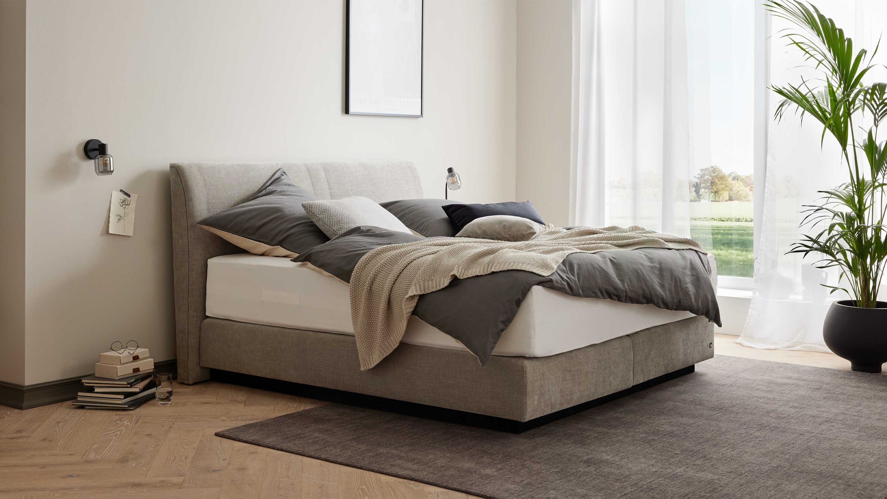 Boxspringbett mit gepolstertem Kopfteil und silberfarbenem Bezug in einem hellen Schlafzimmer