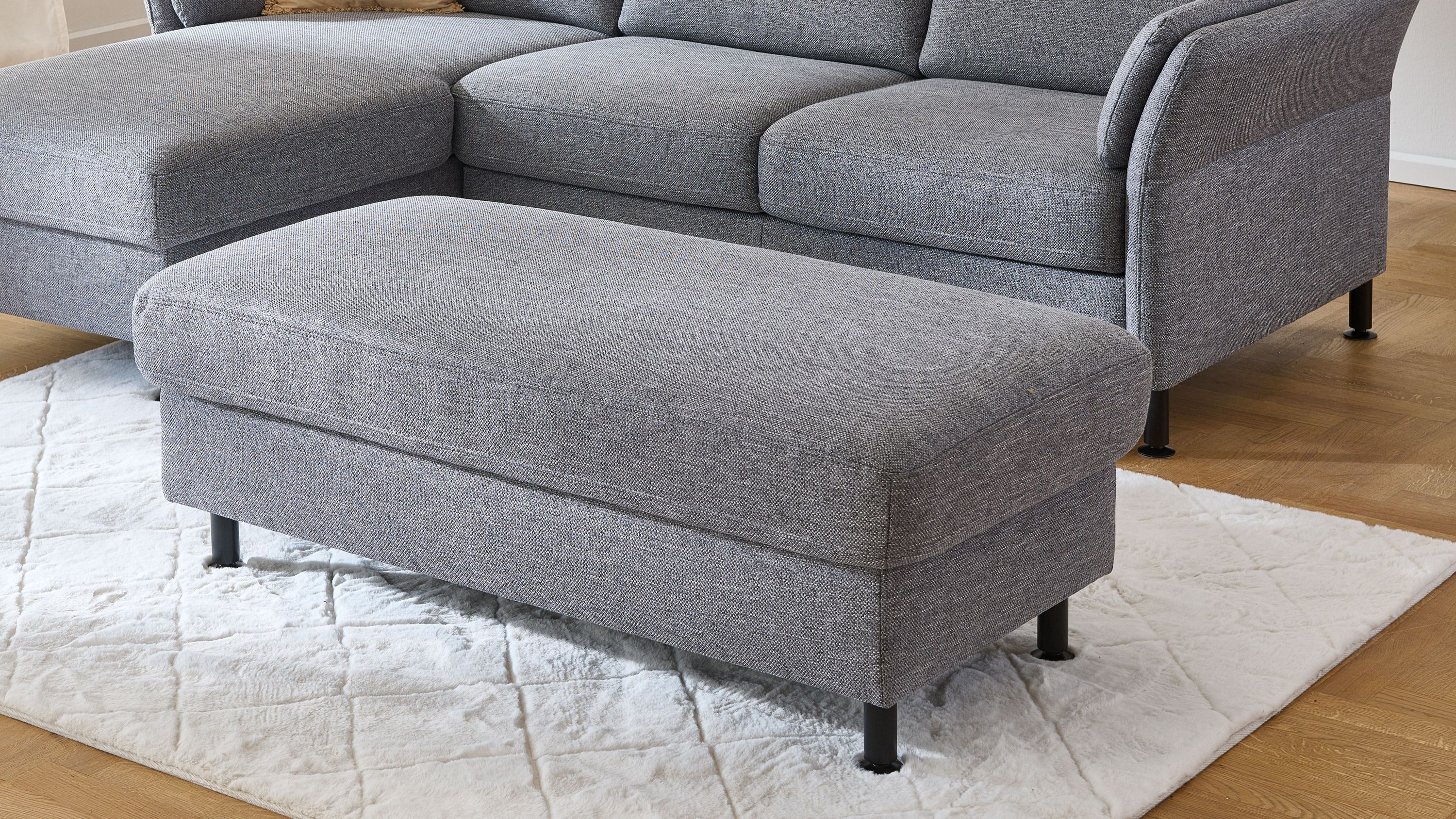 Großer rechteckiger Polsterhocker mit stahlgrauem Flachgewebe und schwarzen Metallfüßen vor grauem Sofa