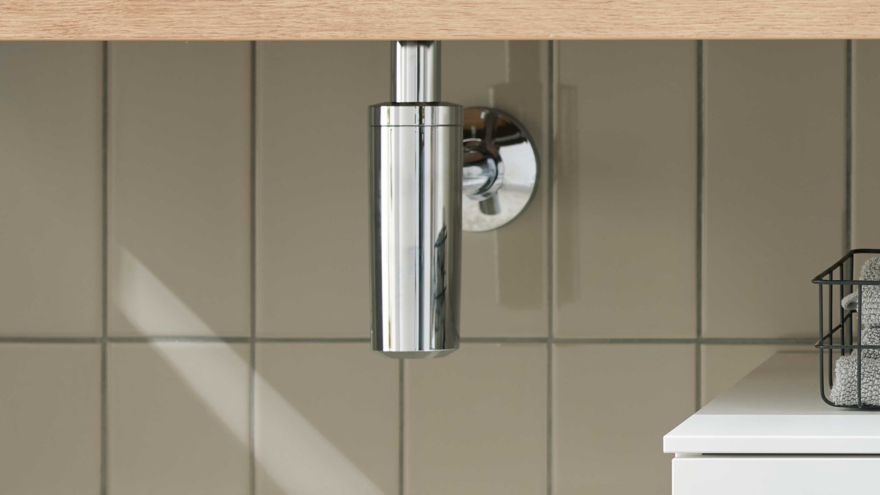 Zylindrischer verchromter Design-Siphon aus Kunststoff unter einem Waschbecken vor beige gekachelter Wand
