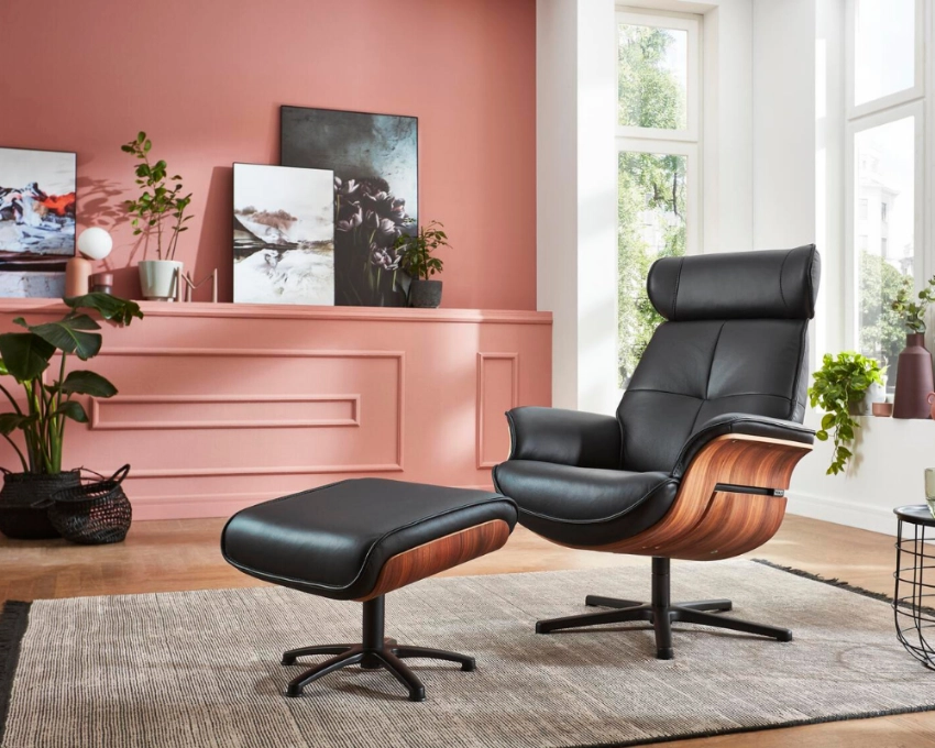Design-Wohnzimmer mit schwarzem Ledersessel und stilvoller Dekoration Schwarzer Ledersessel mit Hocker auf Teppich vor rosa Wand, umgeben von Pflanzen und moderner Wohnzimmereinrichtung