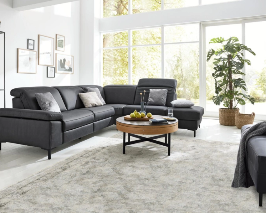 Modernes Wohnzimmer mit grauem Ecksofa, hellen Akzenten und Panoramablick durch bodentiefe Fenster. Wohnzimmer mit grauem Ecksofa, Couchtisch aus Holz und Metall, großem Fenster, Wandbildern und Teppich in Naturtönen