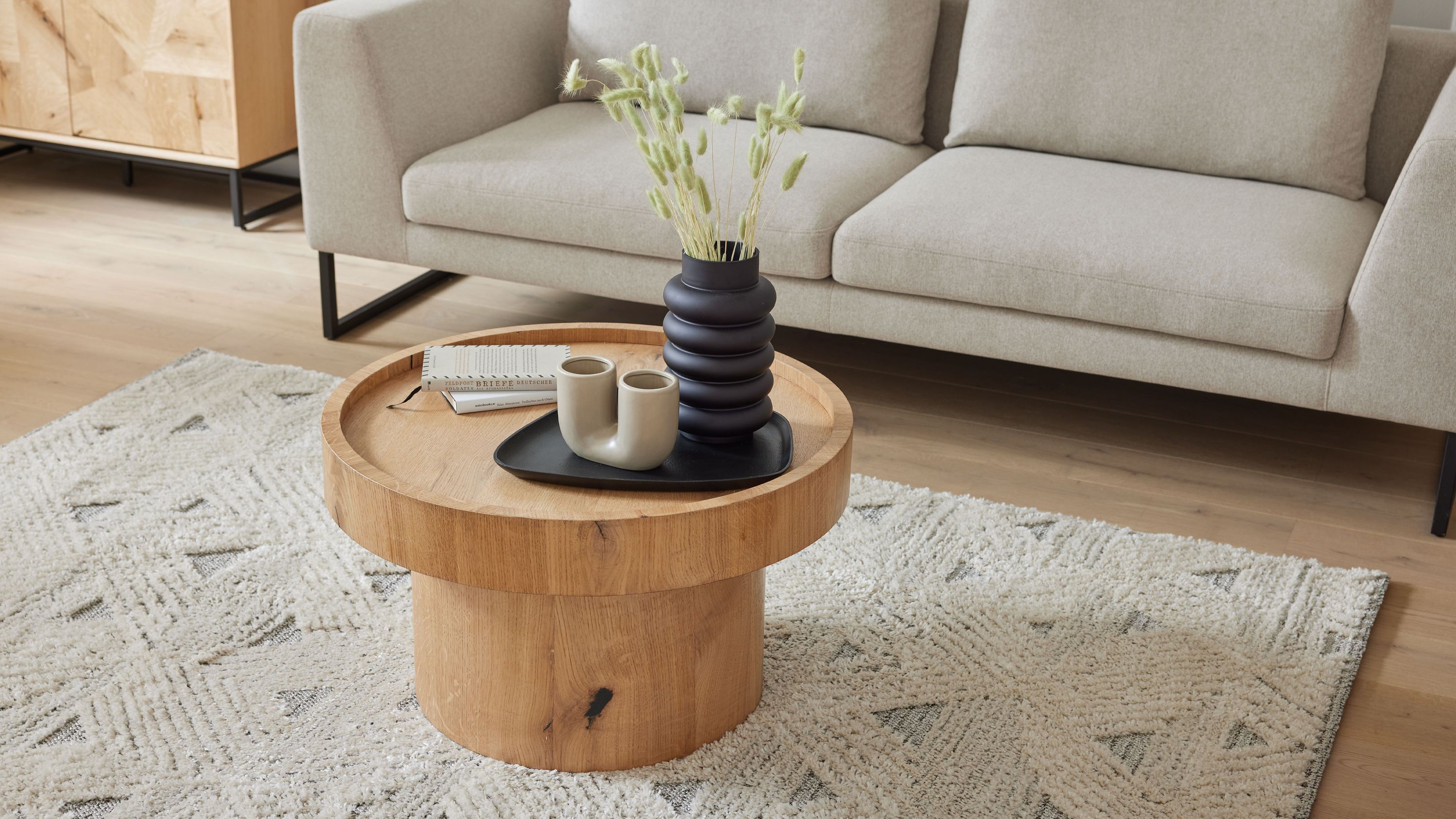 Runder Couchtisch aus hell gewaschenem Holz mit dicker Tischplatte und zylindrischem Fuß auf hellem Teppich vor grauem Sofa