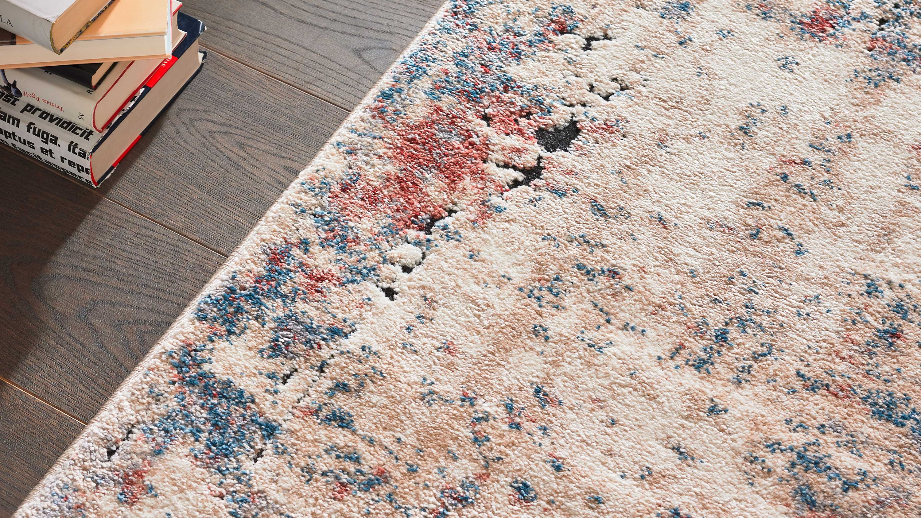 Teppich mit cremefarbenem Grund und roten sowie blauen Mustern auf Holzboden neben Bücherstapel