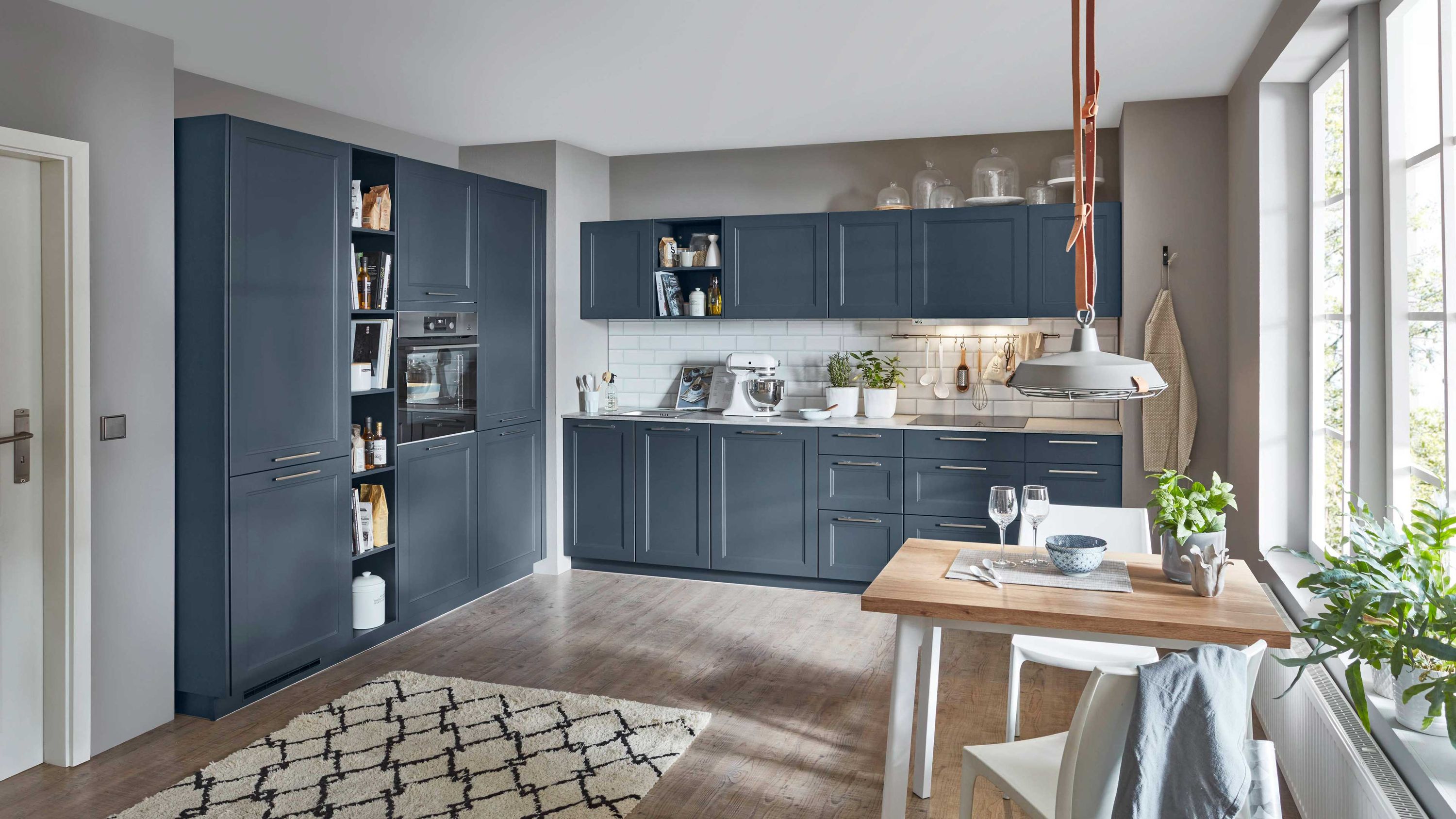 Dunkelblaue Küche mit Holzfußboden Esstisch offenem Regal Einbauofen Teppich Pflanzen Tageslicht.