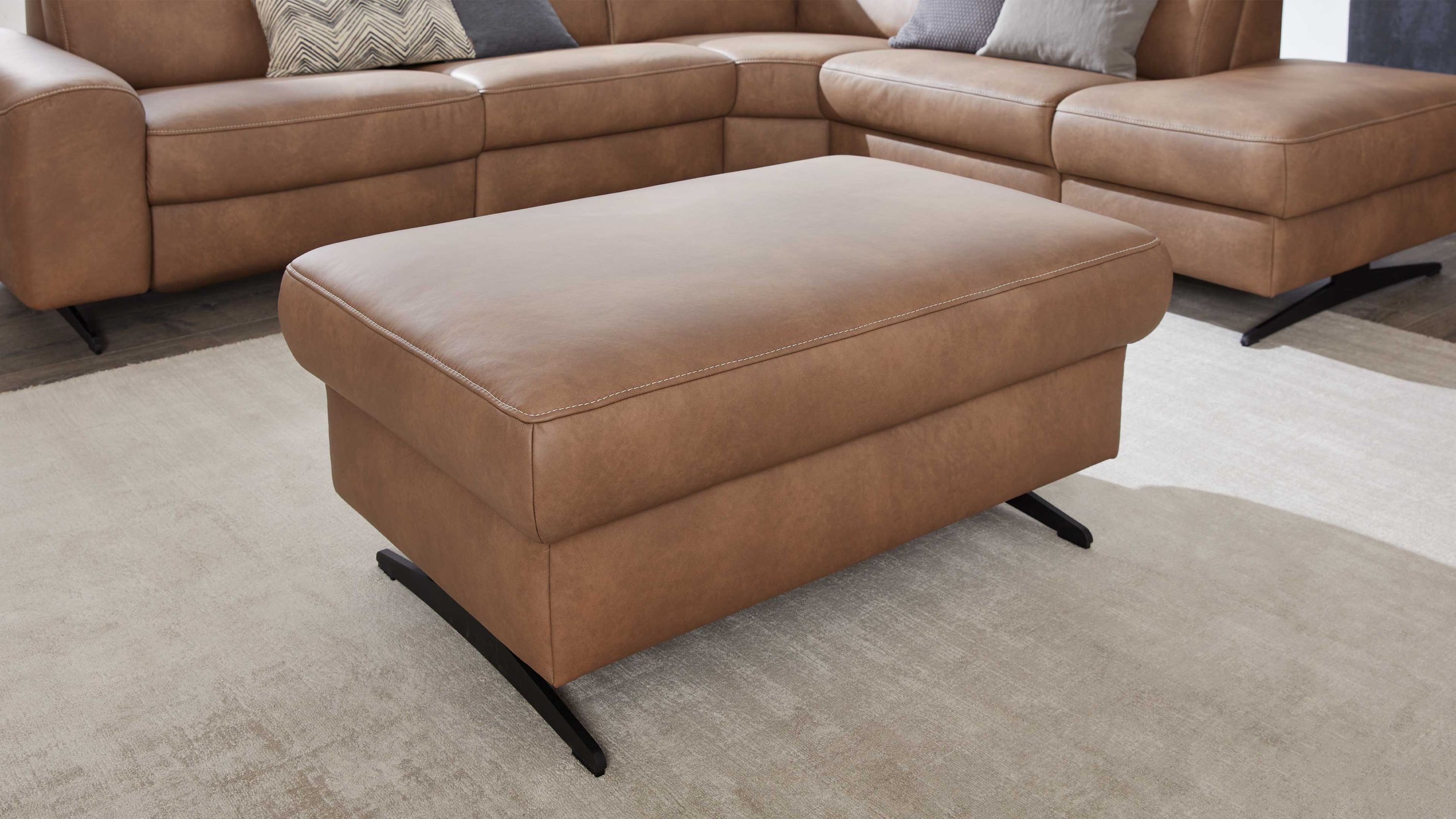 Rechteckiger Hocker in wüstenfarbener Polsterung mit schwarzen Metallfüßen vor braunem Sofa