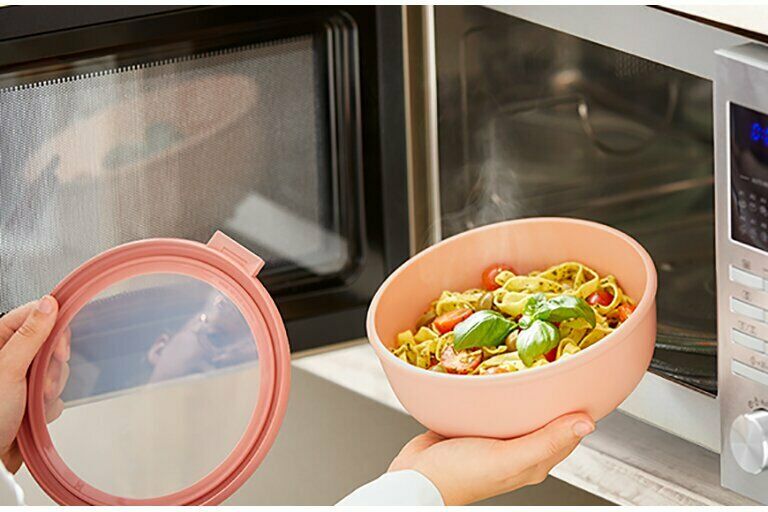 Geöffnete Vorratsdose mit frischem Essen wird vor geöffneter Mikrowelle gehalten  Deckel in der linken Hand sichtbar