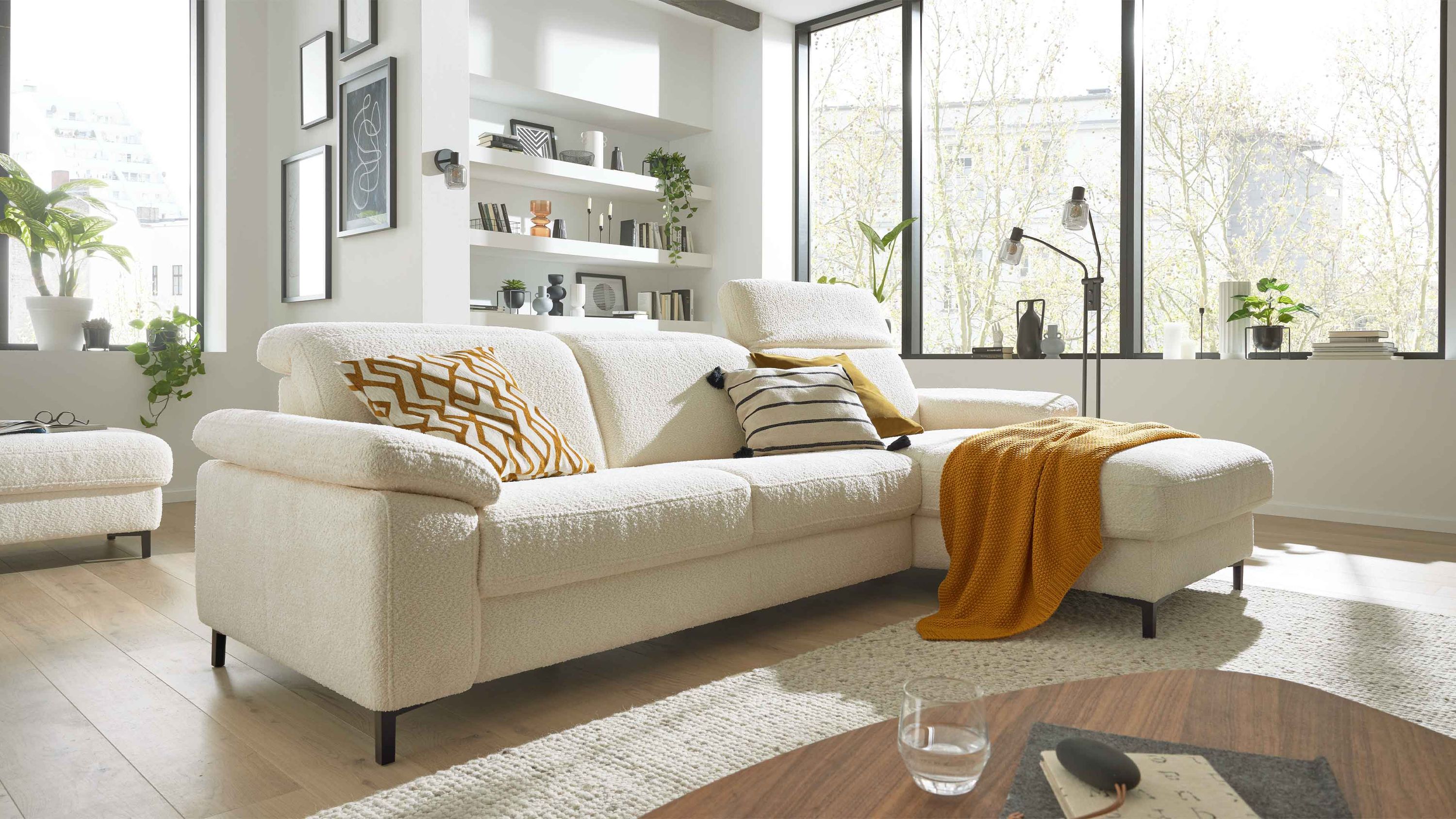 Ecksofa mit naturweißem Chenillebezug schwarzen Metallfüßen mehreren Kissen und einer gelben Decke in hellem Wohnzimmer