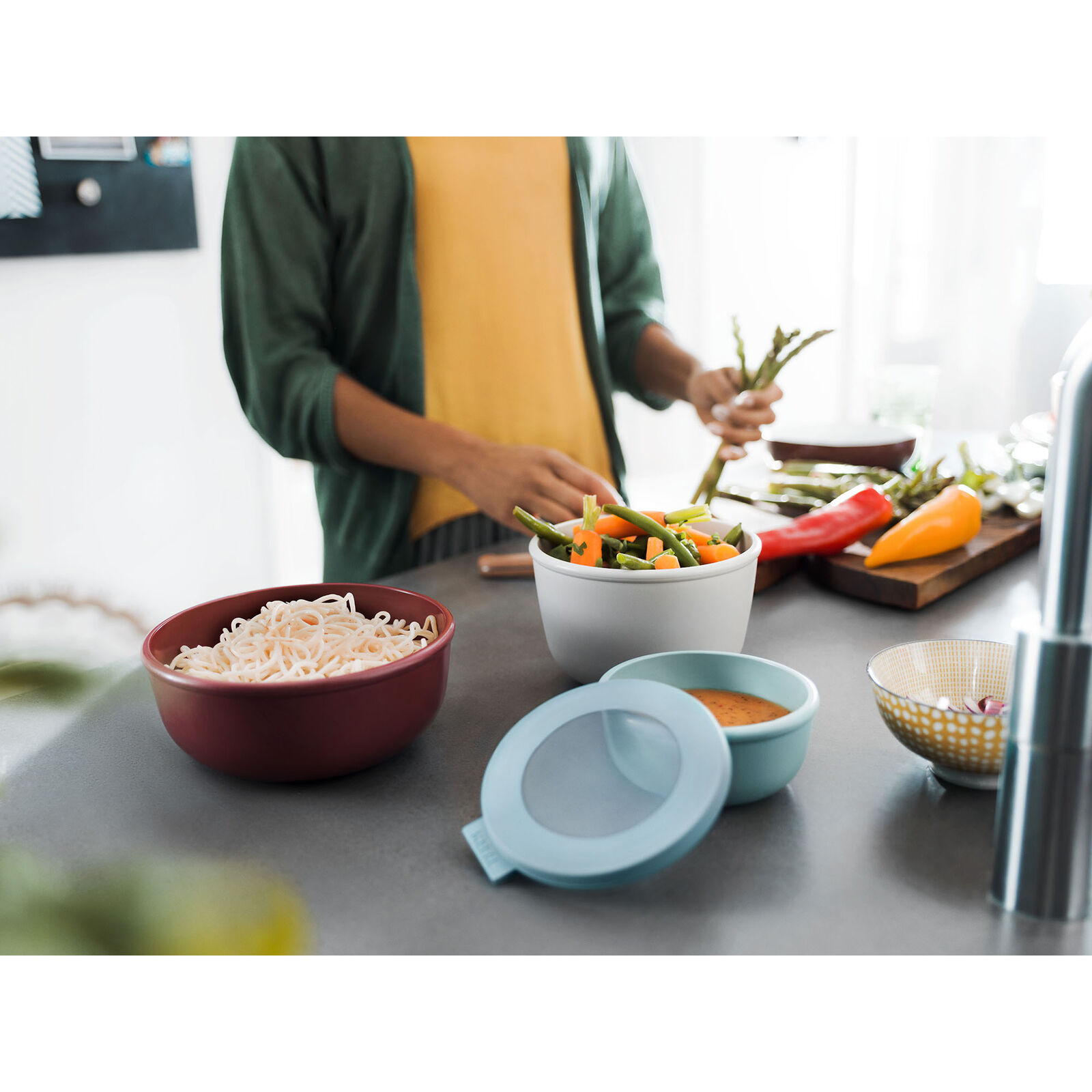 Person bereitet Mahlzeit in moderner Küche zu, verschiedene Multischüsseln mit Zutaten stehen auf Arbeitsfläche