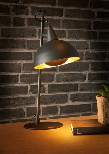 Moderne schwarze Schreibtischlampe auf Holzoberfläche vor Ziegelwand, aus seitlicher Perspektive fotografiert.