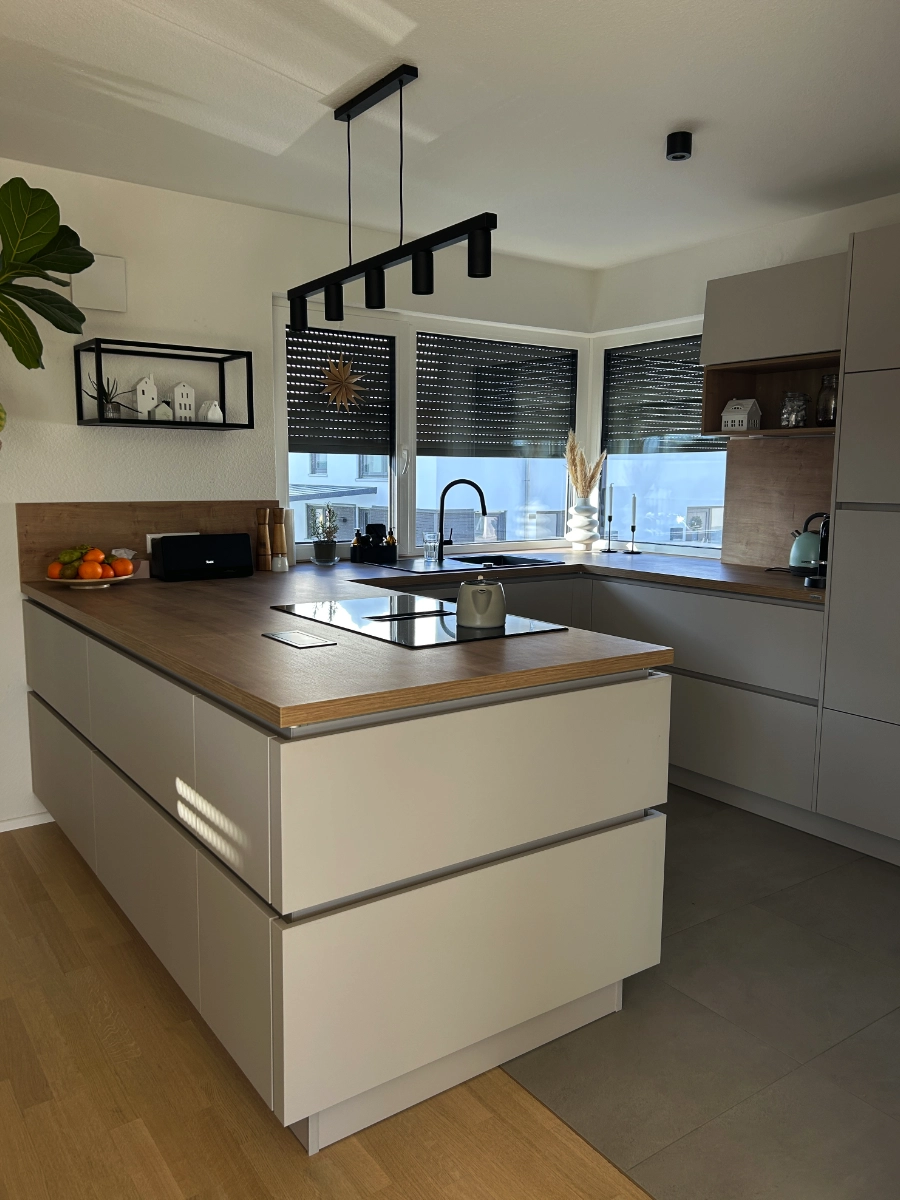 Moderne Wohnküche mit grauer Kochinsel, Holzarbeitsplatte, Induktionskochfeld, Spüle, Hängelampe und großen Fenstern.