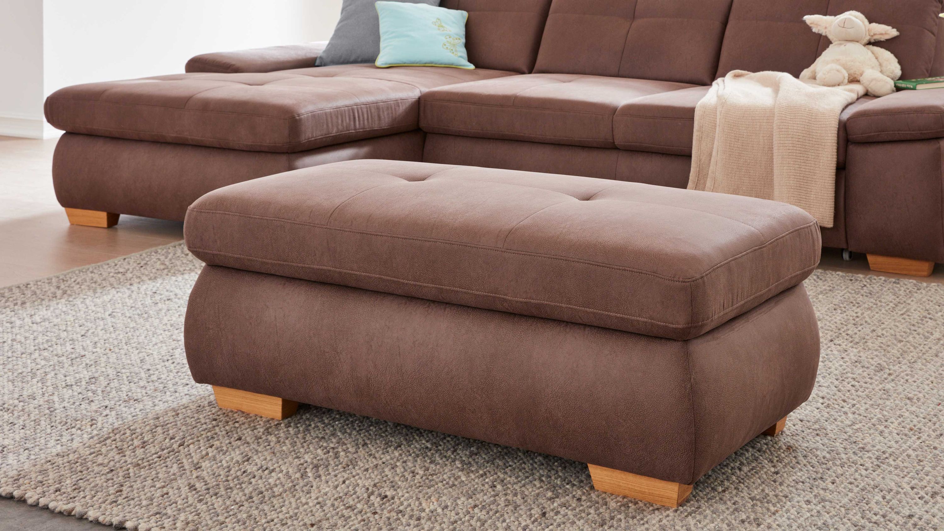 Rechteckiger Hocker mit gepolstertem Sitz in brauner Mikrofaser und kurzen Holzfüßen auf grauem Teppich vor braunem Sofa