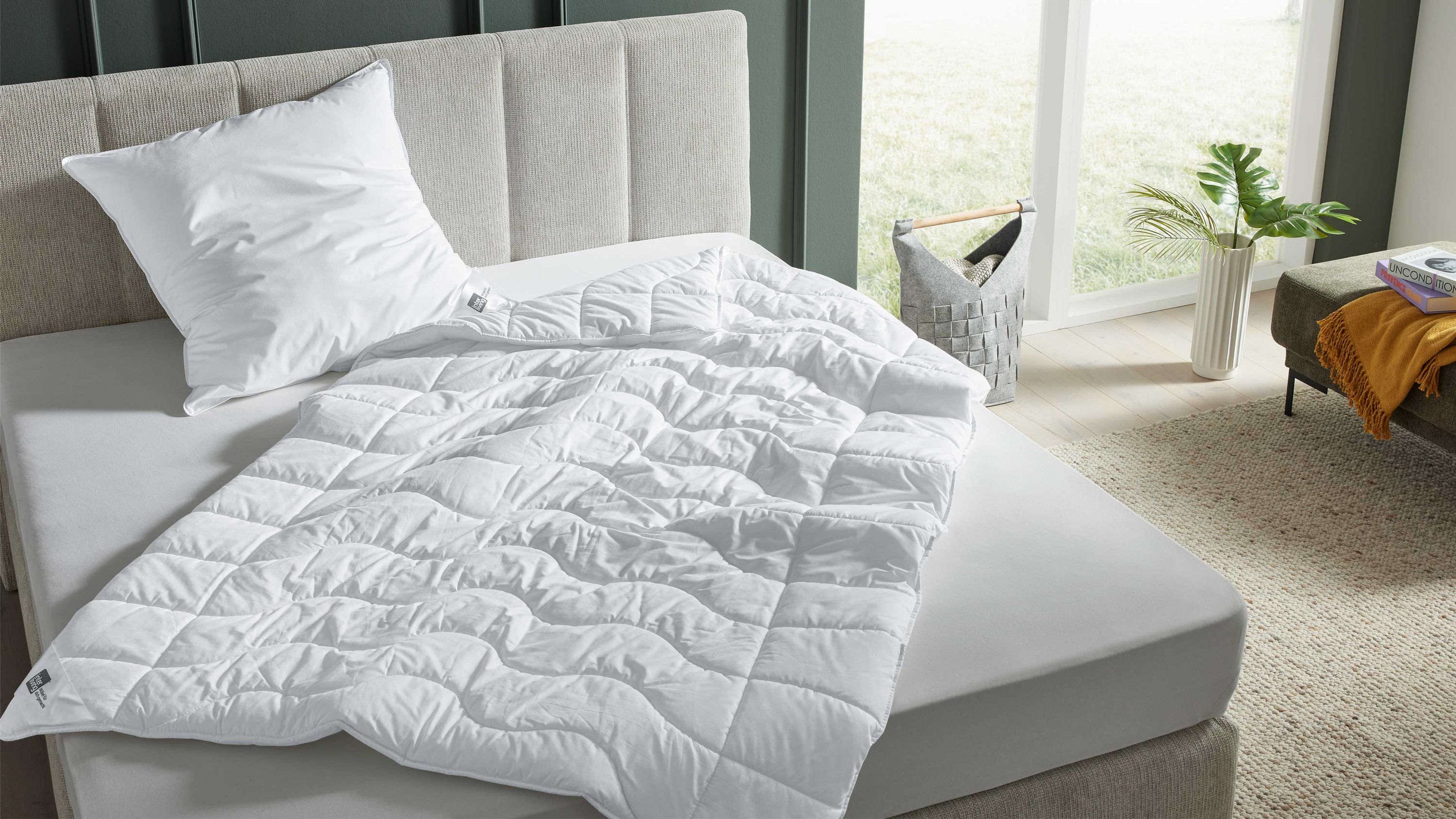 Beiges Bett mit gepolstertem Kopfteil weißem Kissen und weißer Steppdecke in einem hellen Schlafzimmer