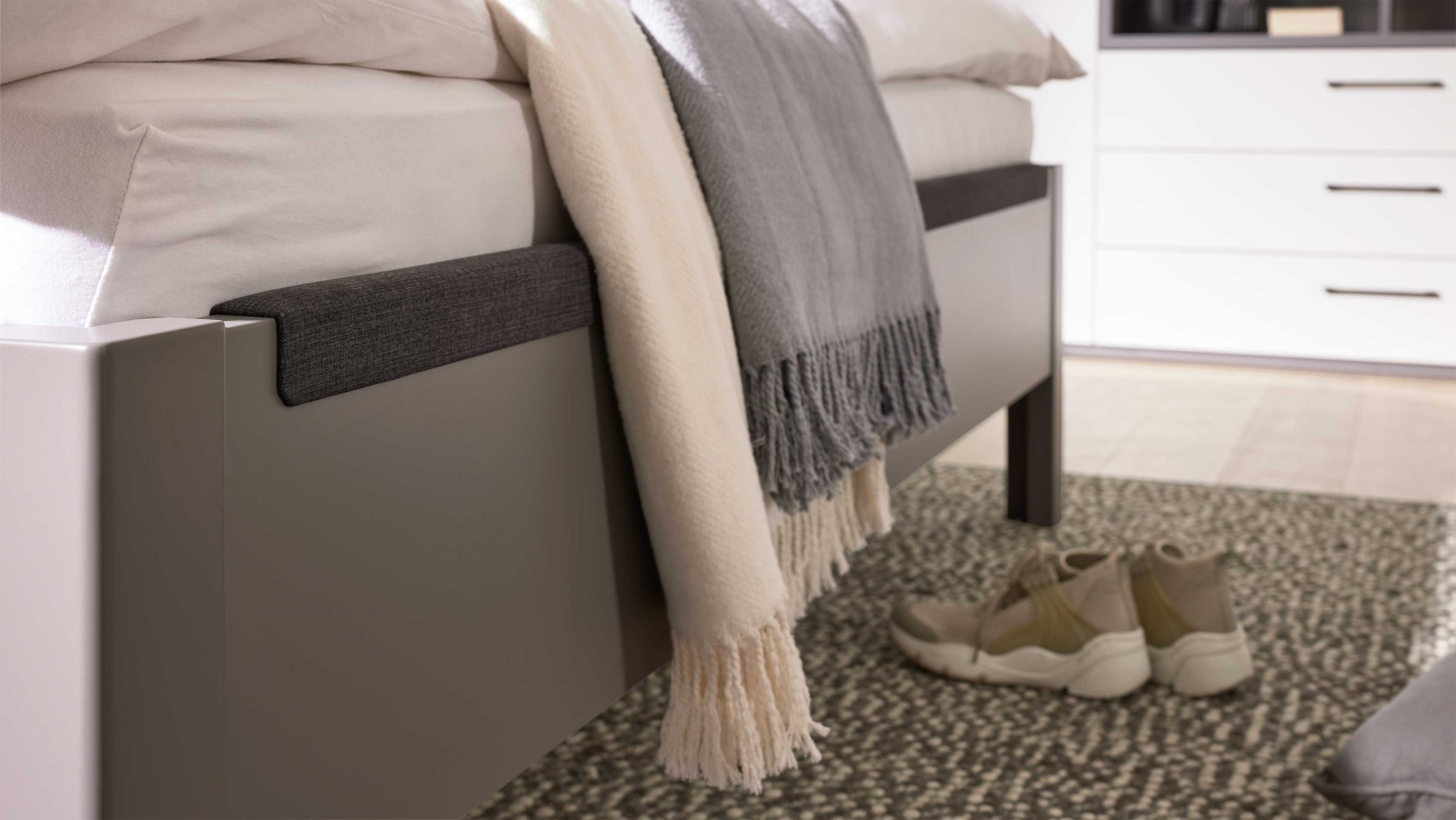 Bettfuß mit anthrazitfarbenem Stoffbezug und zwei Decken in beige und grau auf gemustertem Teppich