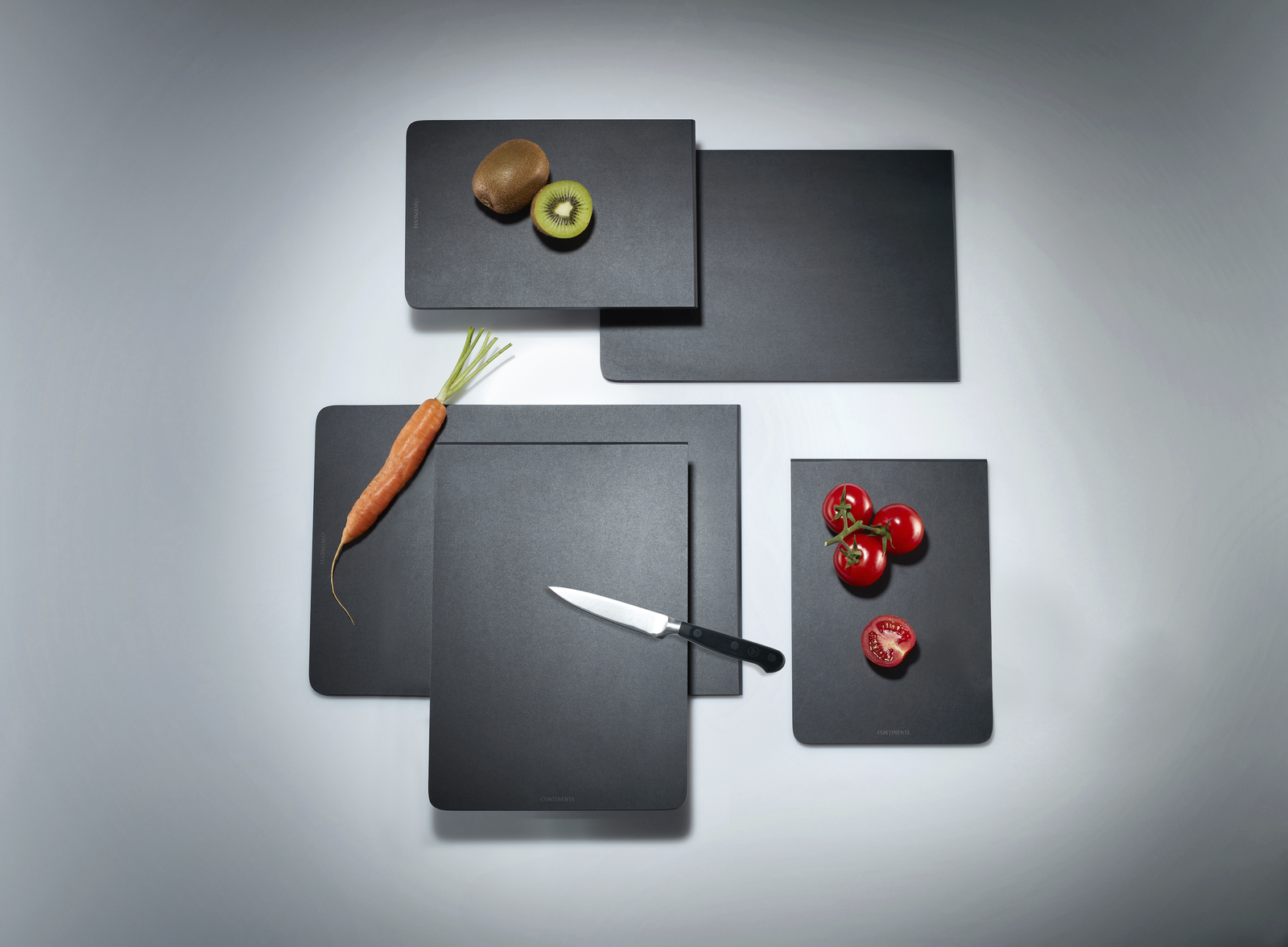 Vier schwarze Schneidebretter mit Kiwi, Karotte, Messer und Tomaten, in gestaffelter Anordnung von oben fotografiert