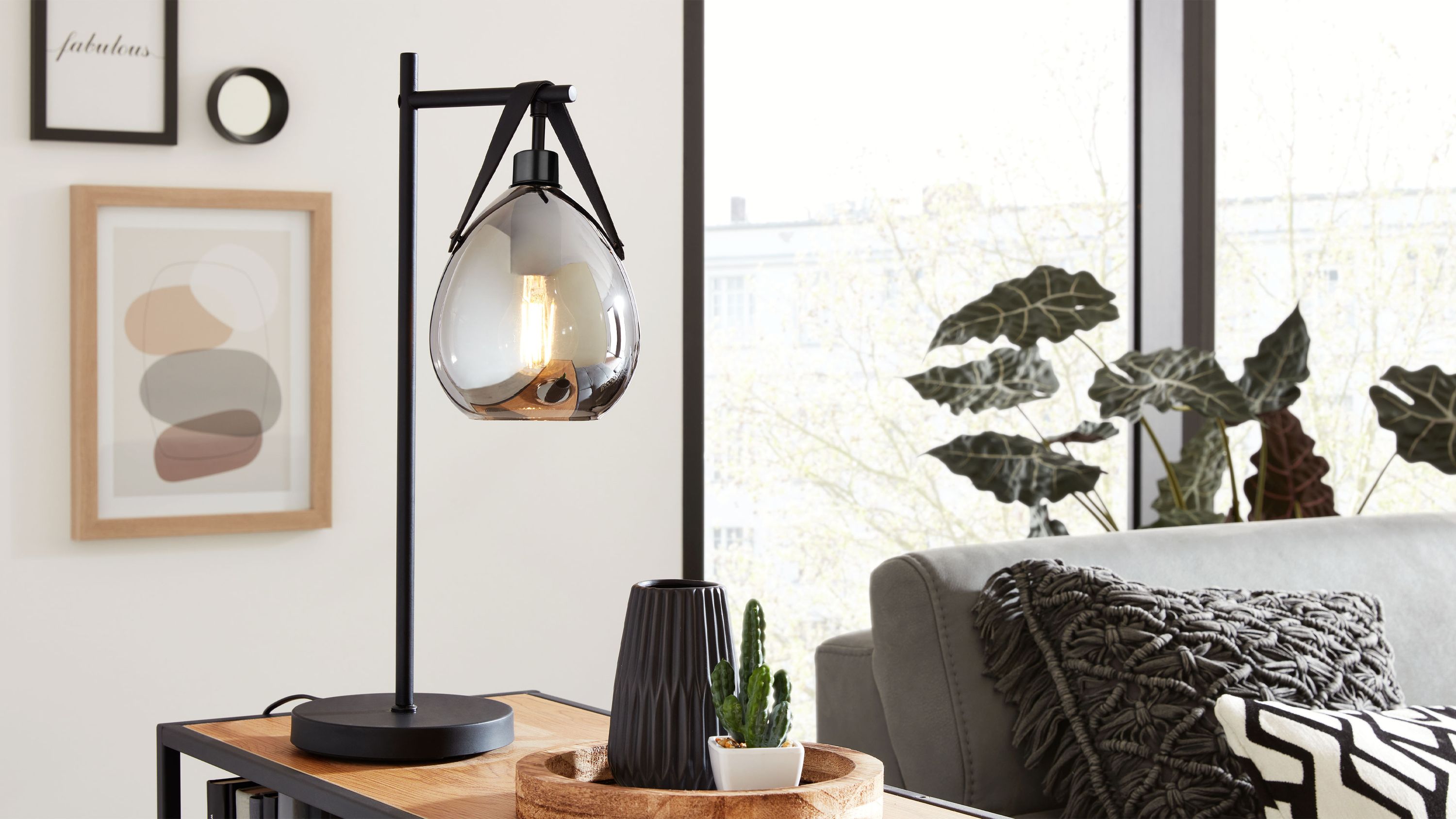 Tischleuchte mit Rauchglas und mattschwarzem Gestell Tischleuchte mit mattschwarzem Gestell und tropfenförmigem Rauchglaslampenschirm auf Holztisch neben grauem Sofa