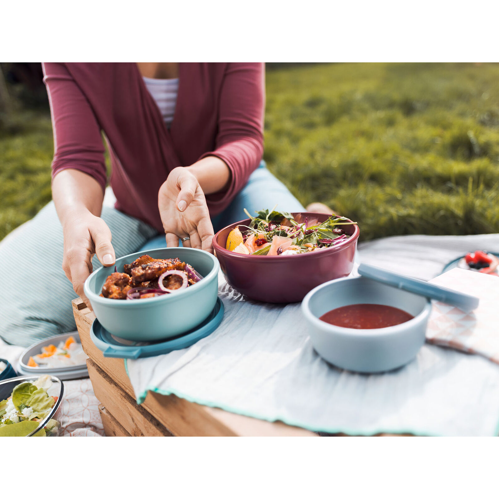 Picknick auf einer Wiese mit verschiedenen gefüllten Vorratsdosen auf Decke  Person greift nach einer Dose mit Snacks
