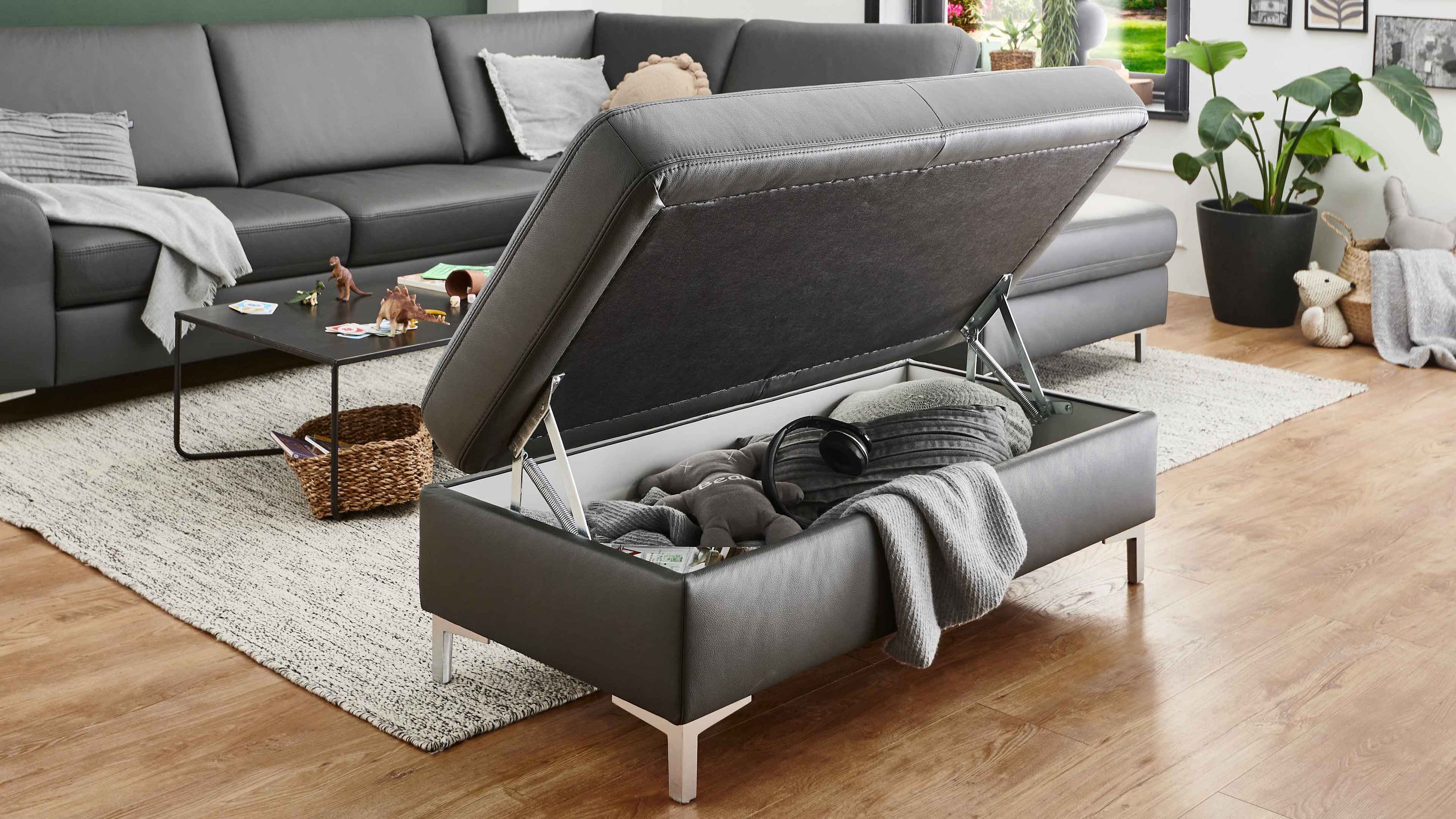 Anthrazitfarbener Hocker mit Stauraum und Alu-Winkelfüßen in einem Wohnzimmer mit grauem Sofa