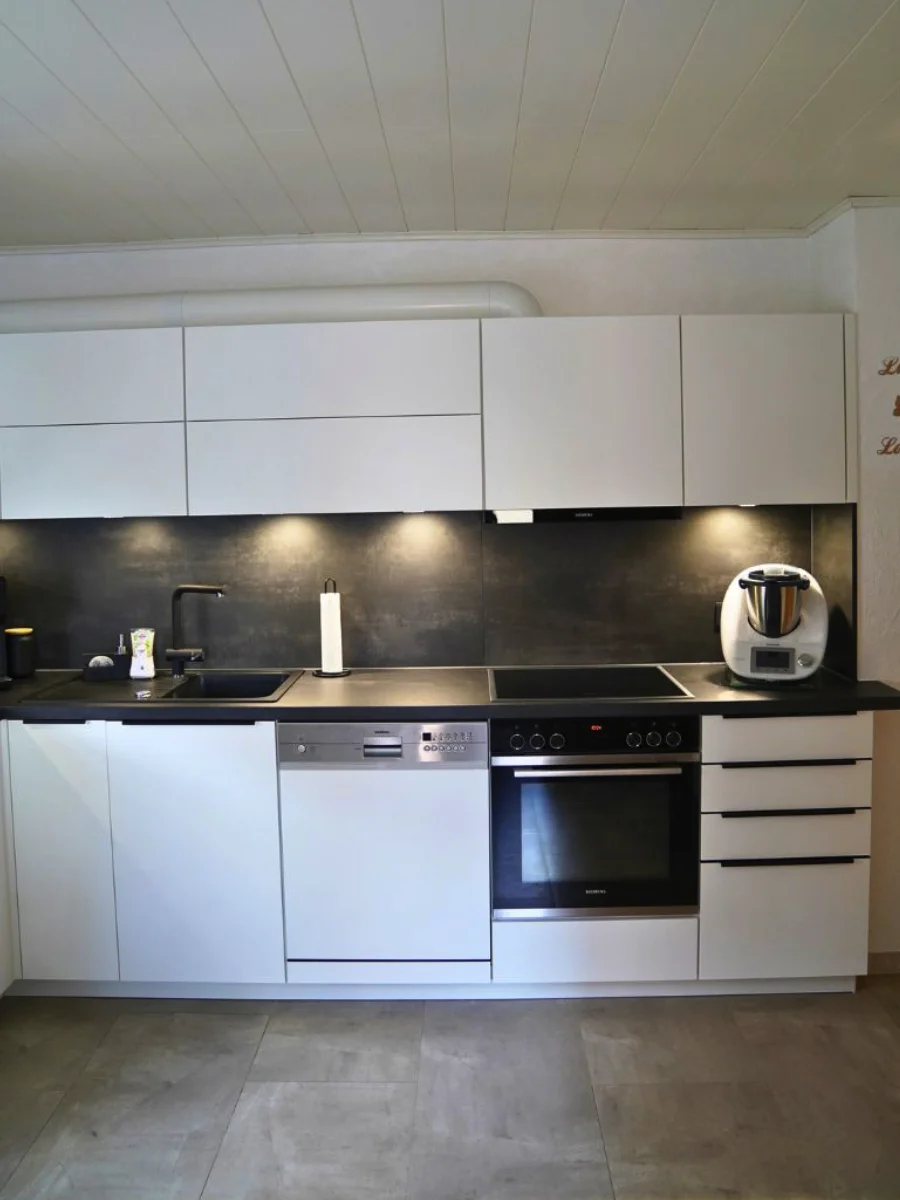 Moderne weiße Eckküche mit schwarzen Arbeitsflächen, Kaffeemaschine, Spüle, Einbaugeräten und Pflanze auf Fliesenboden.