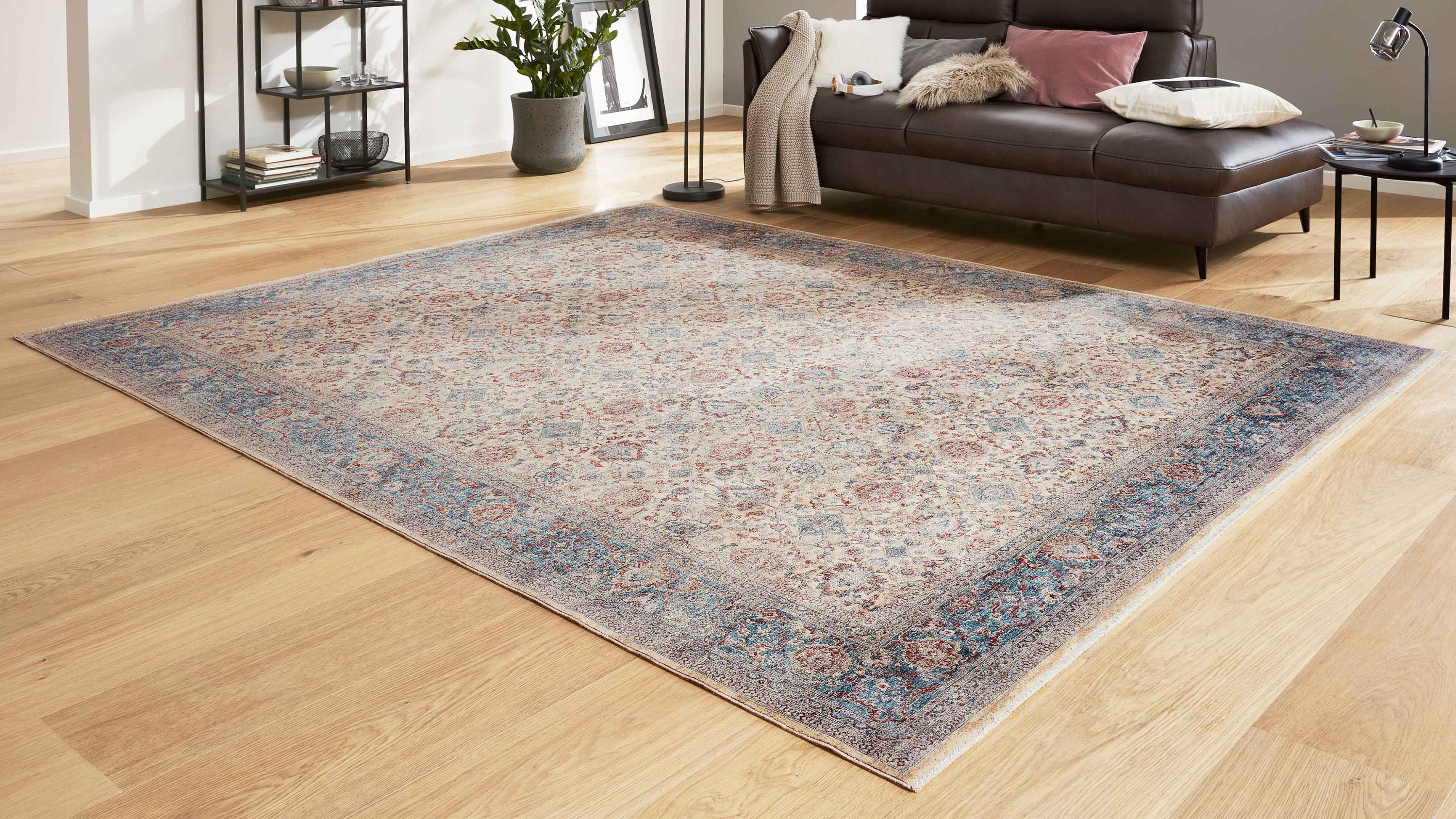Rechteckiger Teppich mit cremefarbenem Grund und blauem Muster auf hellem Holzboden vor braunem Sofa
