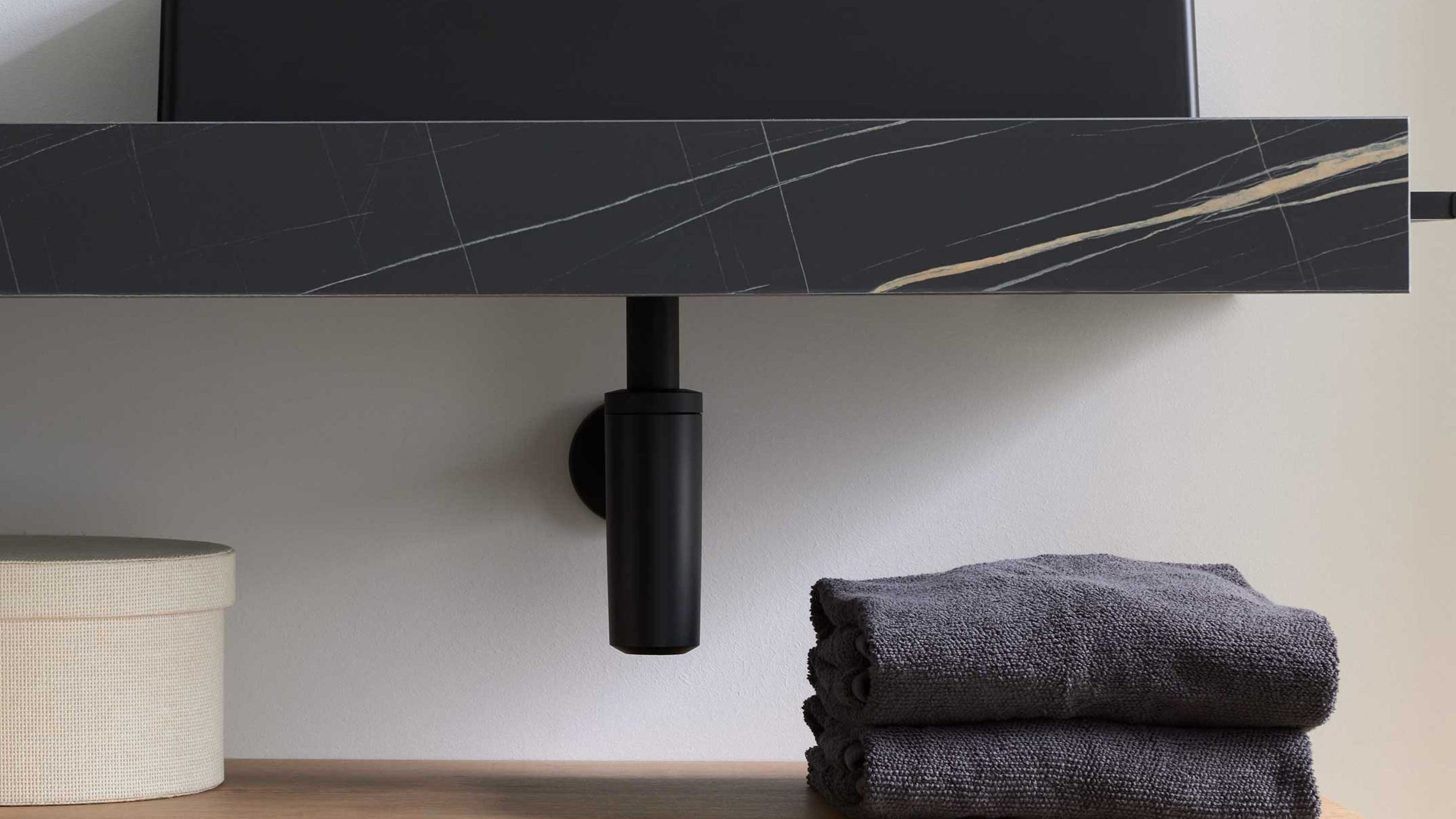 Mattschwarzer Design-Siphon unter einem Waschbecken mit dunkler Oberfläche und grauen Handtüchern daneben