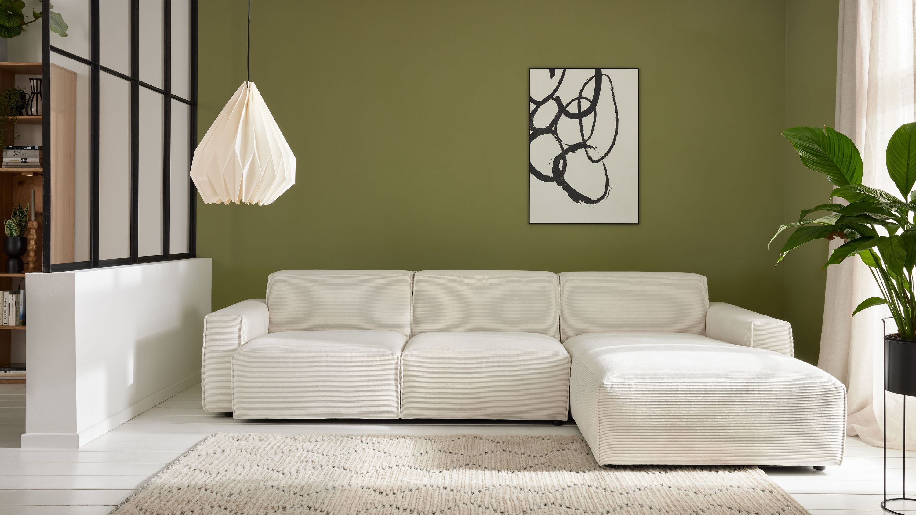 Ecksofa mit drei Sitzflächen und Ottomane in weißem Stoff vor grüner Wand mit abstraktem Bild