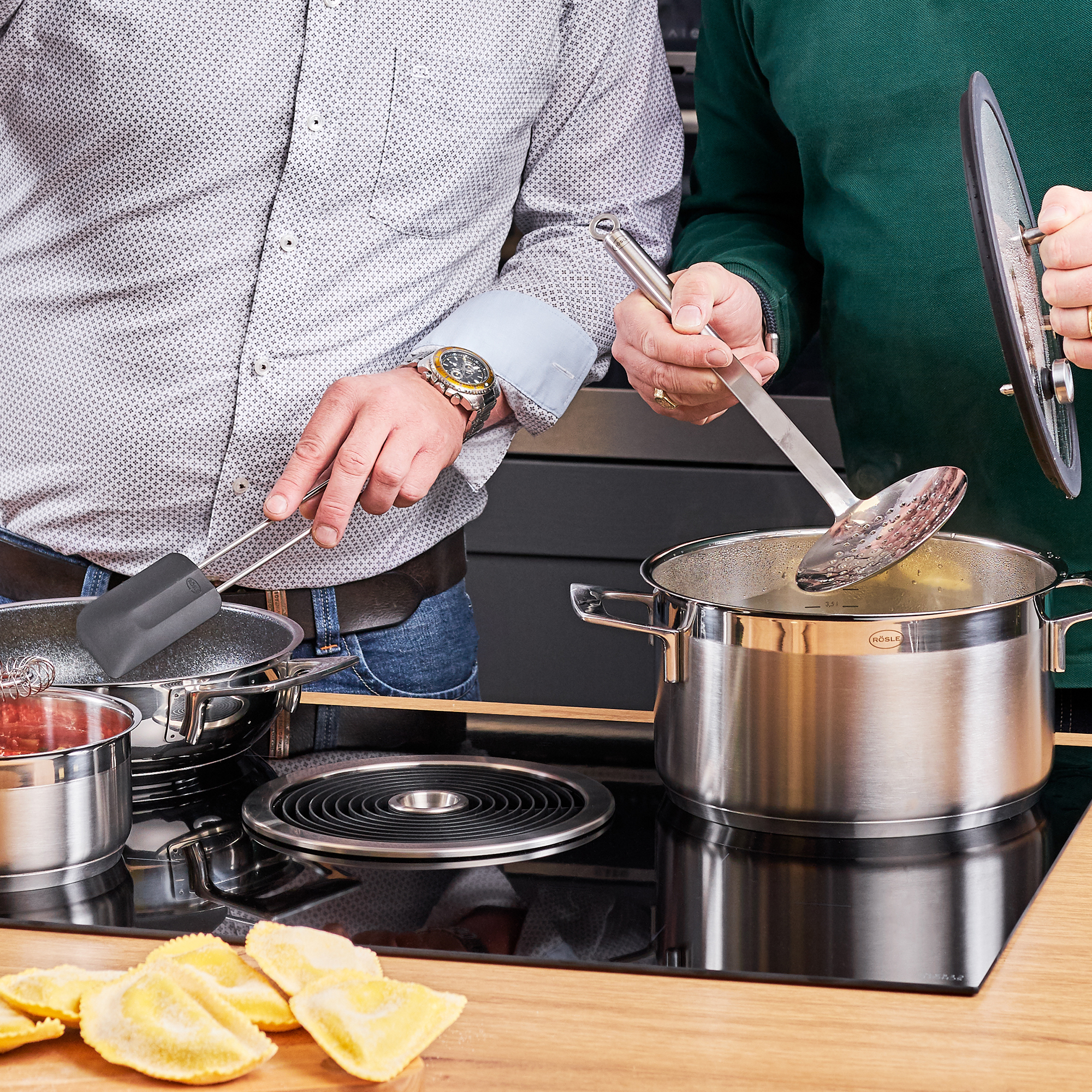 Schaumlöffel aus Edelstahl beim Kochen am Herd daneben Person mit Utensilien schräg von links