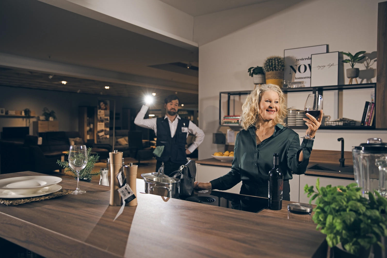 Frau genießt Wein in Küchenausstellung Frau steht in moderner Küchenausstellung, hält ein Glas Rotwein, im Hintergrund steht ein Mitarbeiter mit Taschenlampe.